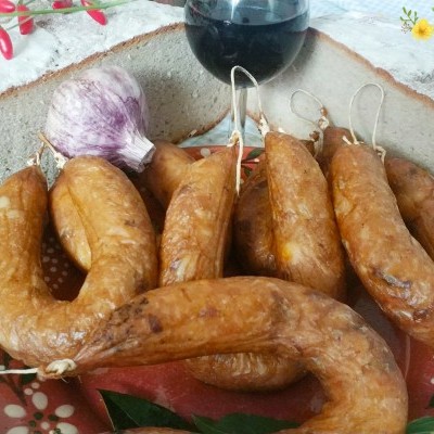 Prato de cerâmica com várias chouriças castanhas, alho roxo e garrafa pequena sobre superfície de madeira e toalha floral branca