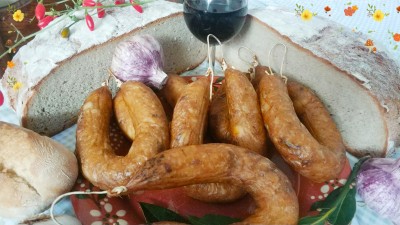 Prato de cerâmica com várias chouriças castanhas, alho roxo e garrafa pequena sobre superfície de madeira e toalha floral branca