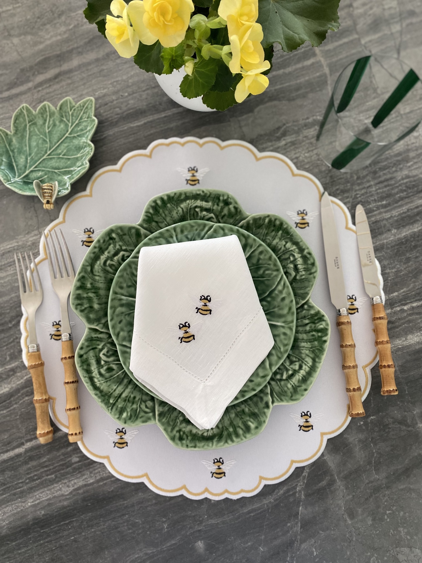 Conjunto de loiça com prato branco decorado com abelhas, prato verde tipo folha, guardanapo branco com abelhas e talheres de cabo madeira