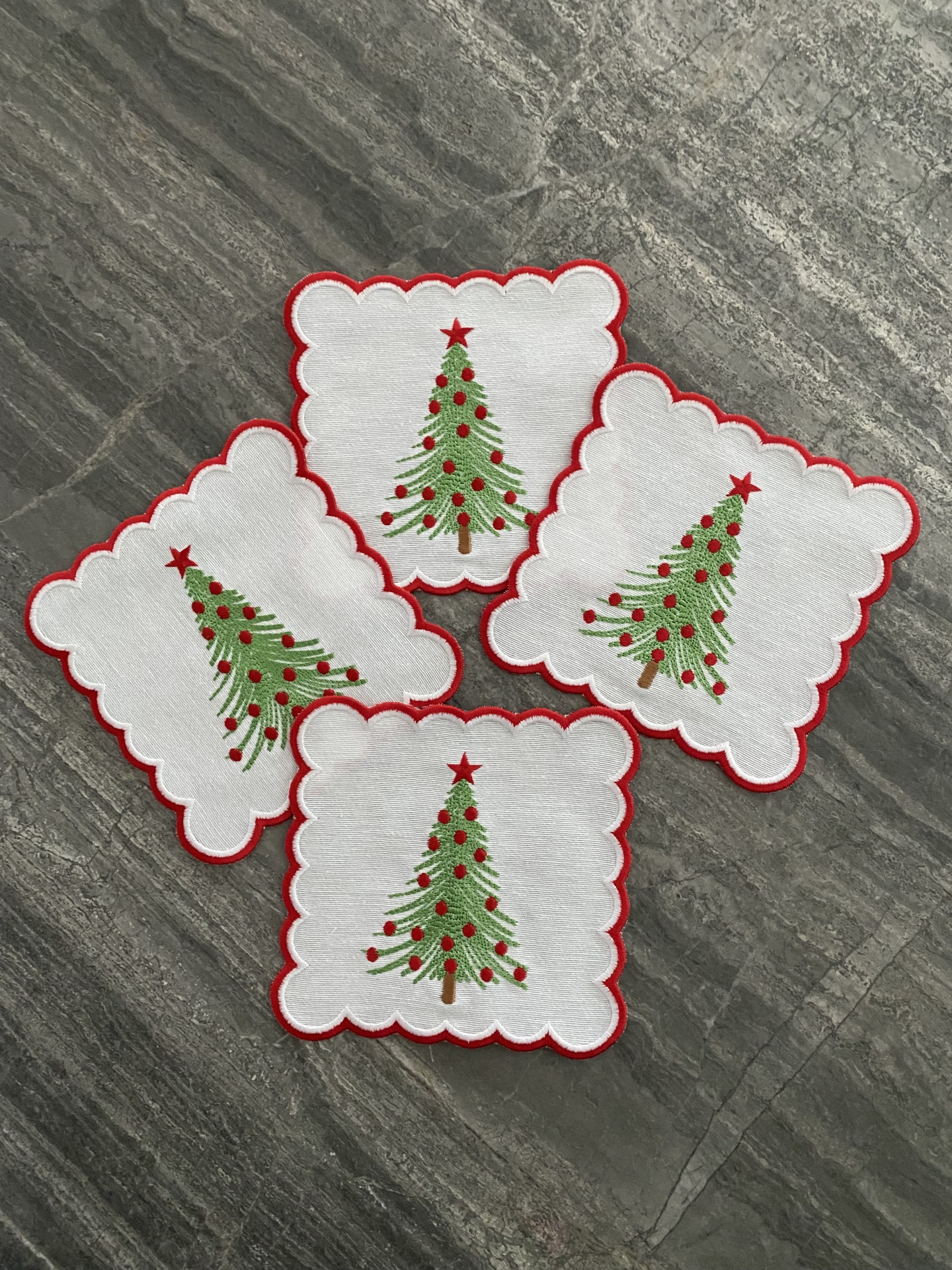 Quatro guardanapos brancos com borda vermelha e árvore de Natal bordada, sobre superfície cinza.