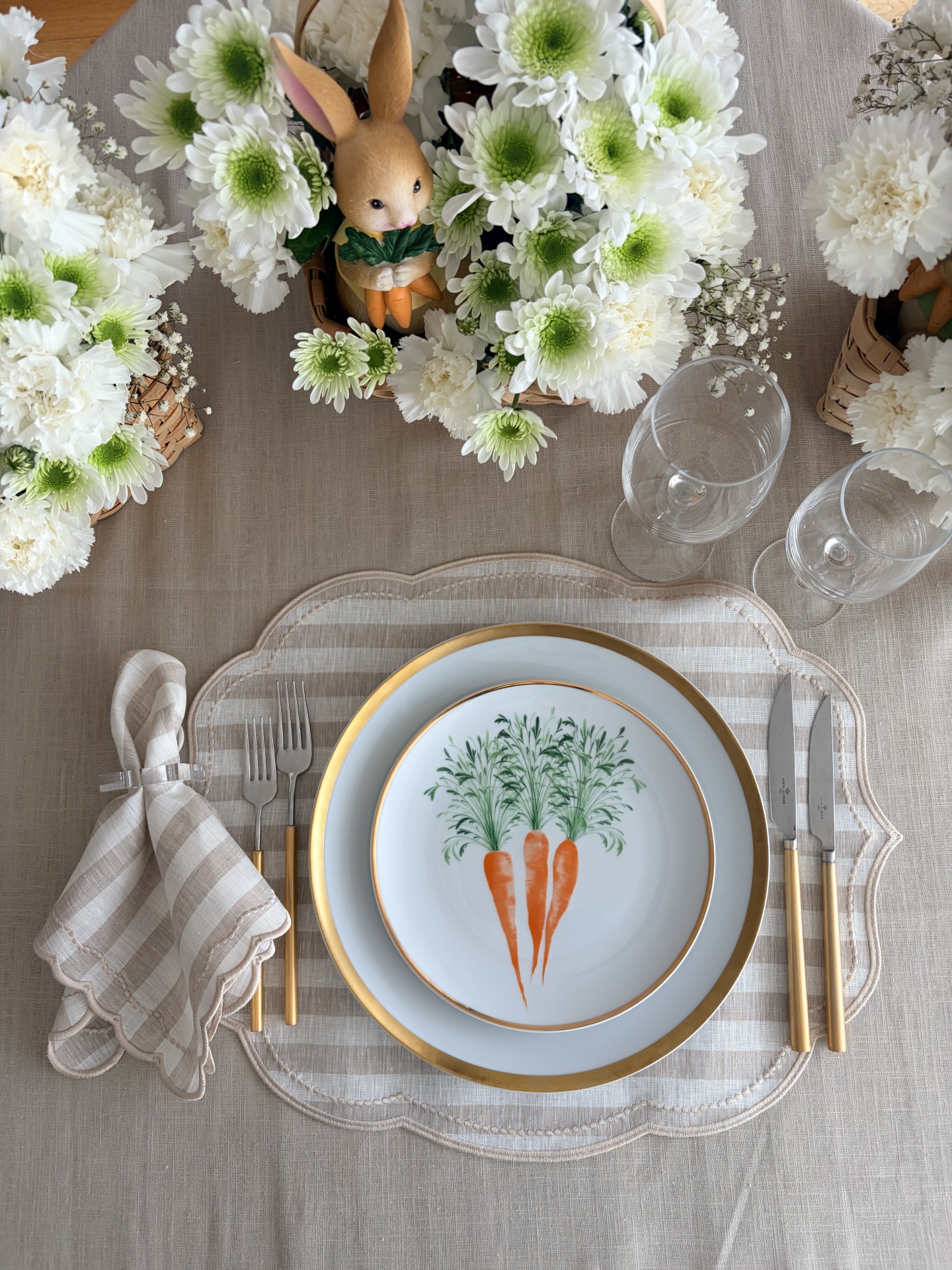 Conjunto de mesa com prato decorado com cenouras, talheres dourados, guardanapo às riscas e decoração com flores e coelho