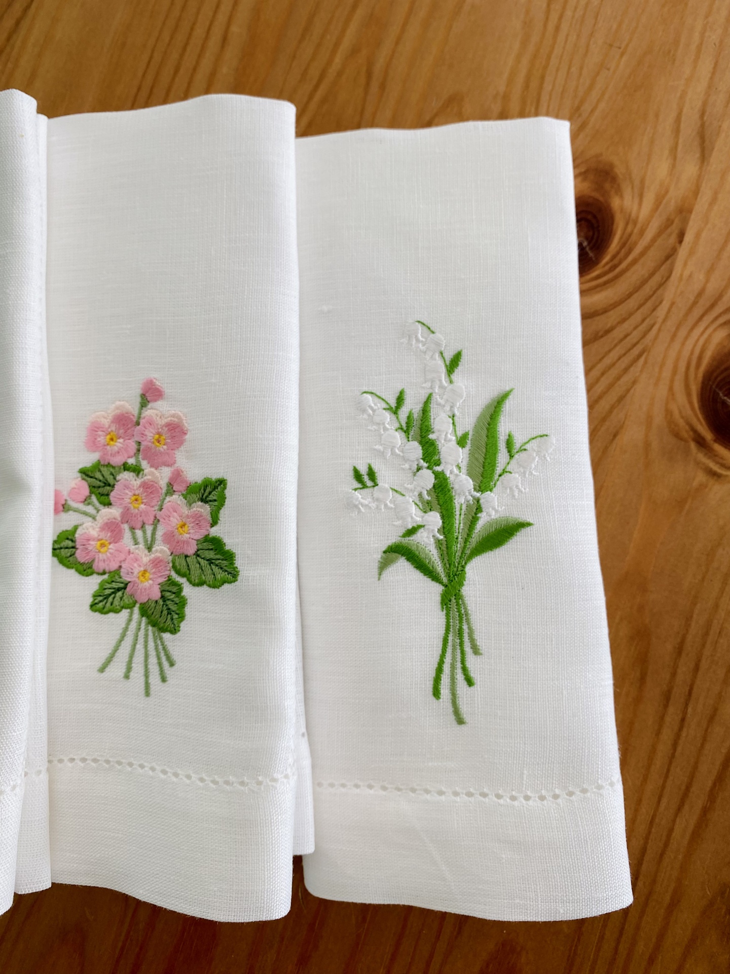 Guardanapos brancos com bordados florais rosa e branco sobre mesa de madeira.