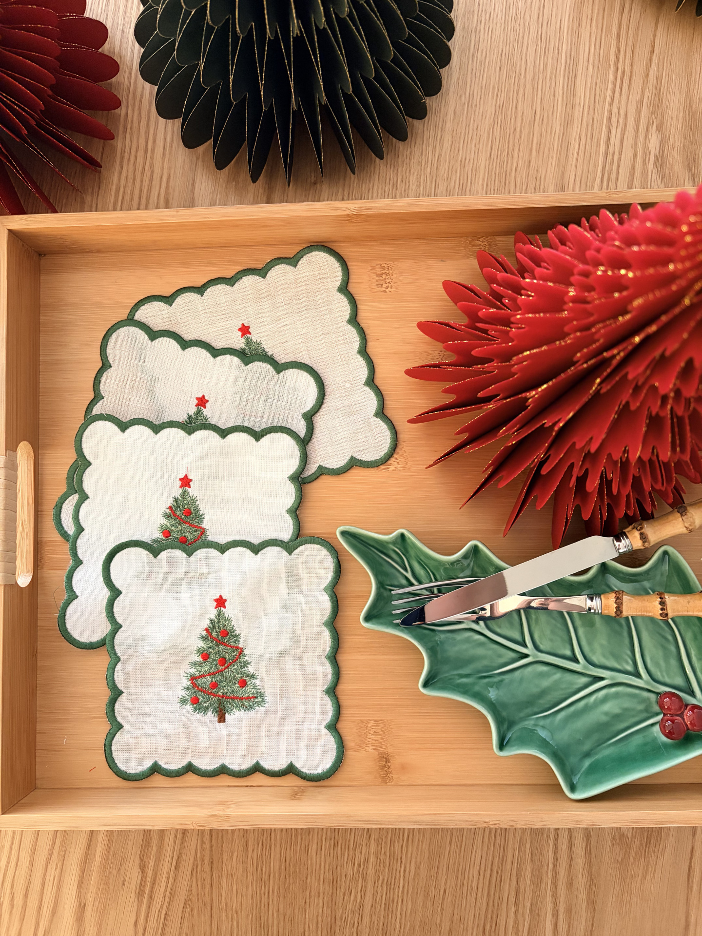 Toalhetes de Natal de linho branco com árvore verde, prato em forma de folha verde com frutas vermelhas e utensílios com cabo bambu numa bandeja de madeira clara.