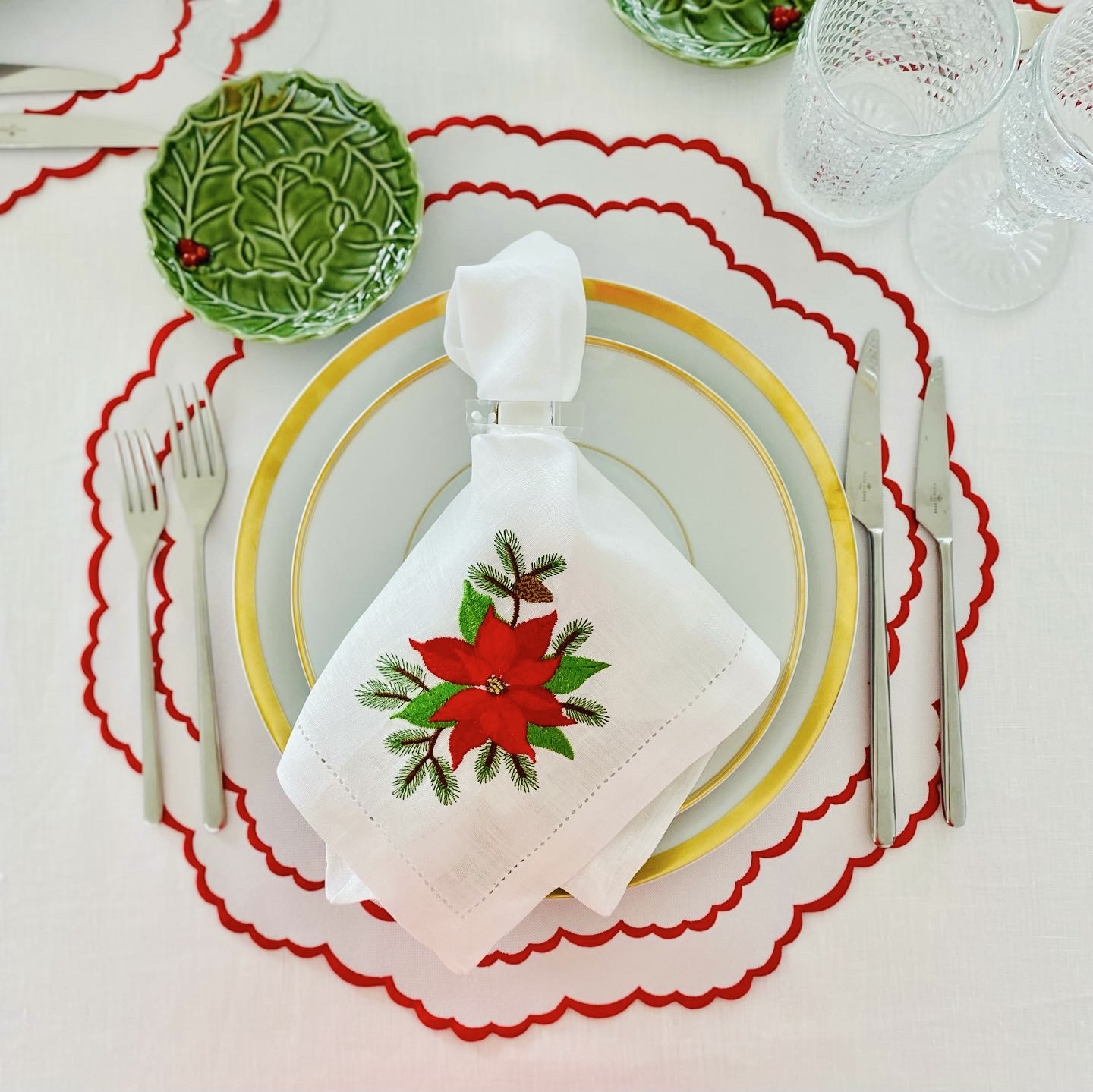 Conjunto de pratos brancos com borda dourada, guardanapo branco bordado em flor vermelha e folhas verdes, pratos verdes em relevo e copos de vidro sobre toalha branca com sousplats brancos e bordas vermelhas
