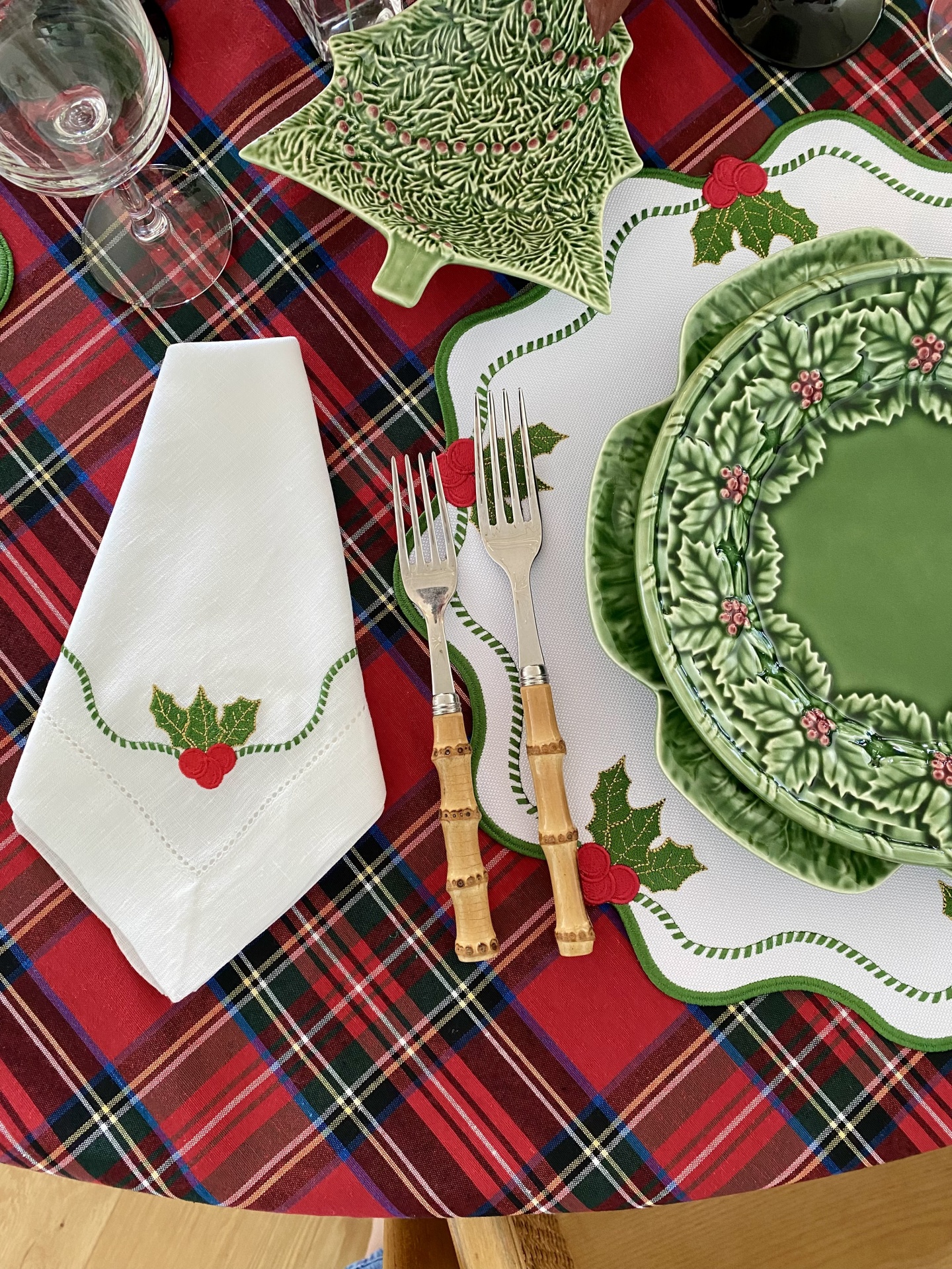 Mesa posta com pratos verdes decorados com folhas de azevinho, talheres com cabo de madeira, guardanapo branco com bordado de azevinho e toalha tartan.