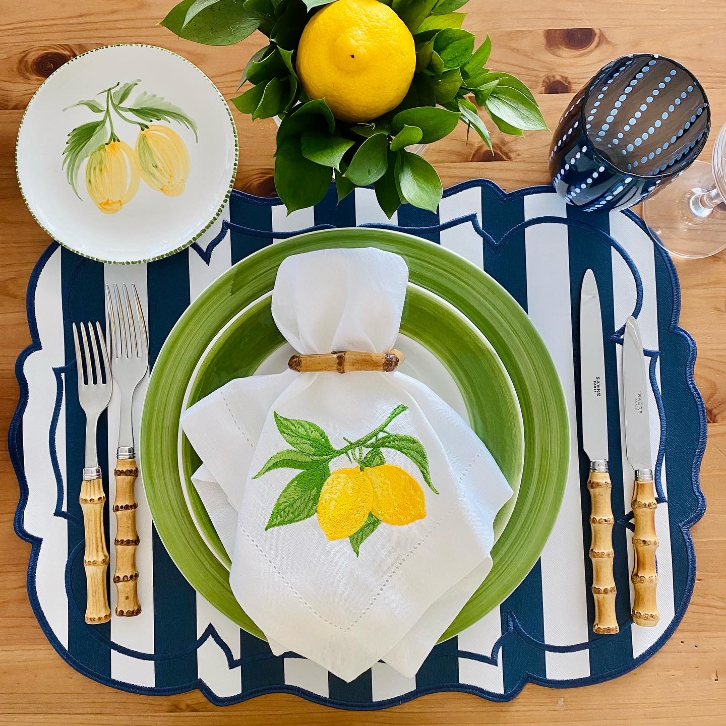 Mesa posta com prato verde, talheres de cabo castanho, prato com limões pintados, guardanapo branco com limões bordados e copo azul de bolinhas
