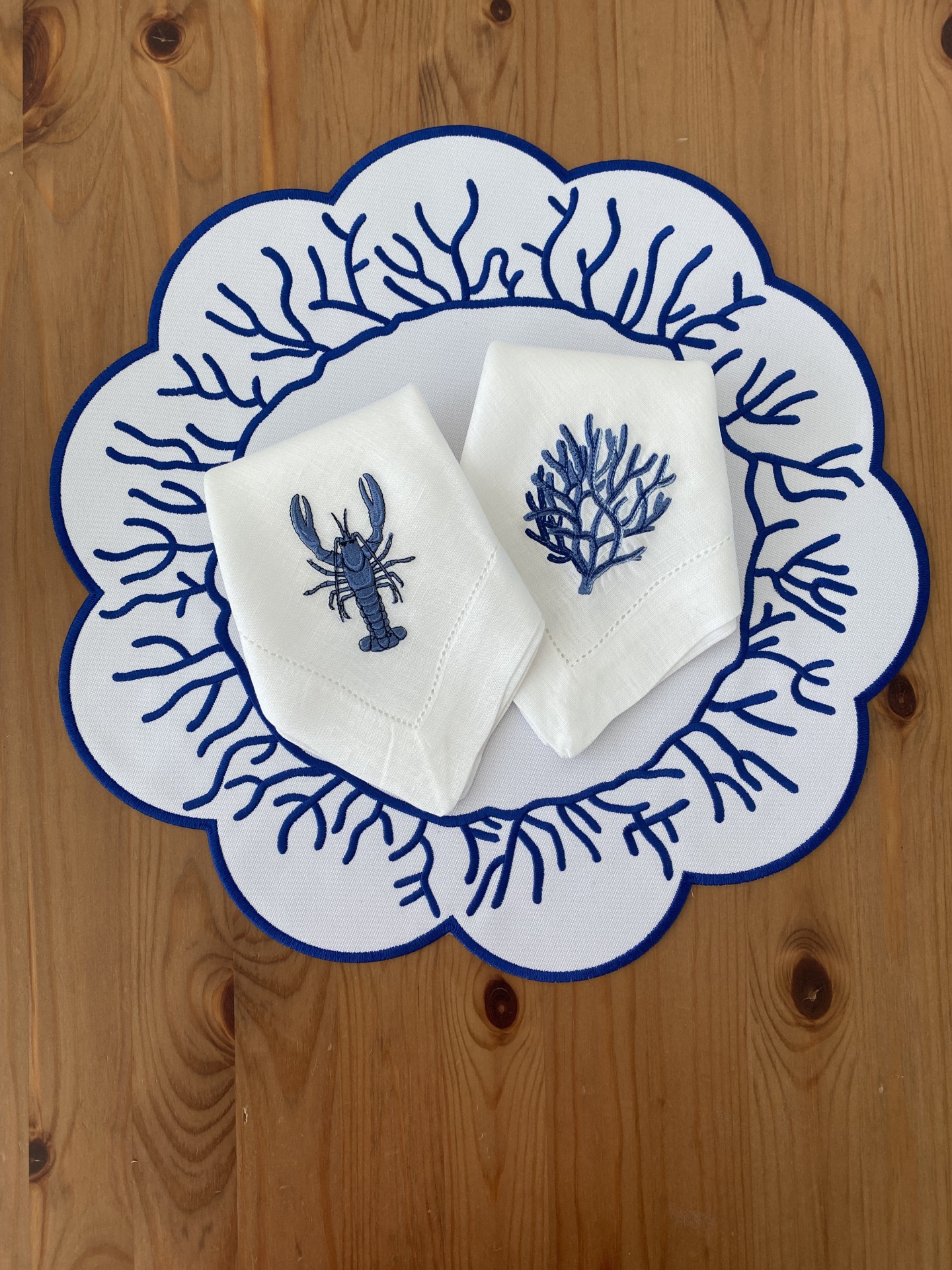 Guardanapos brancos bordados com desenhos azuis de coral e lagostim sobre descanso de prato branco e azul