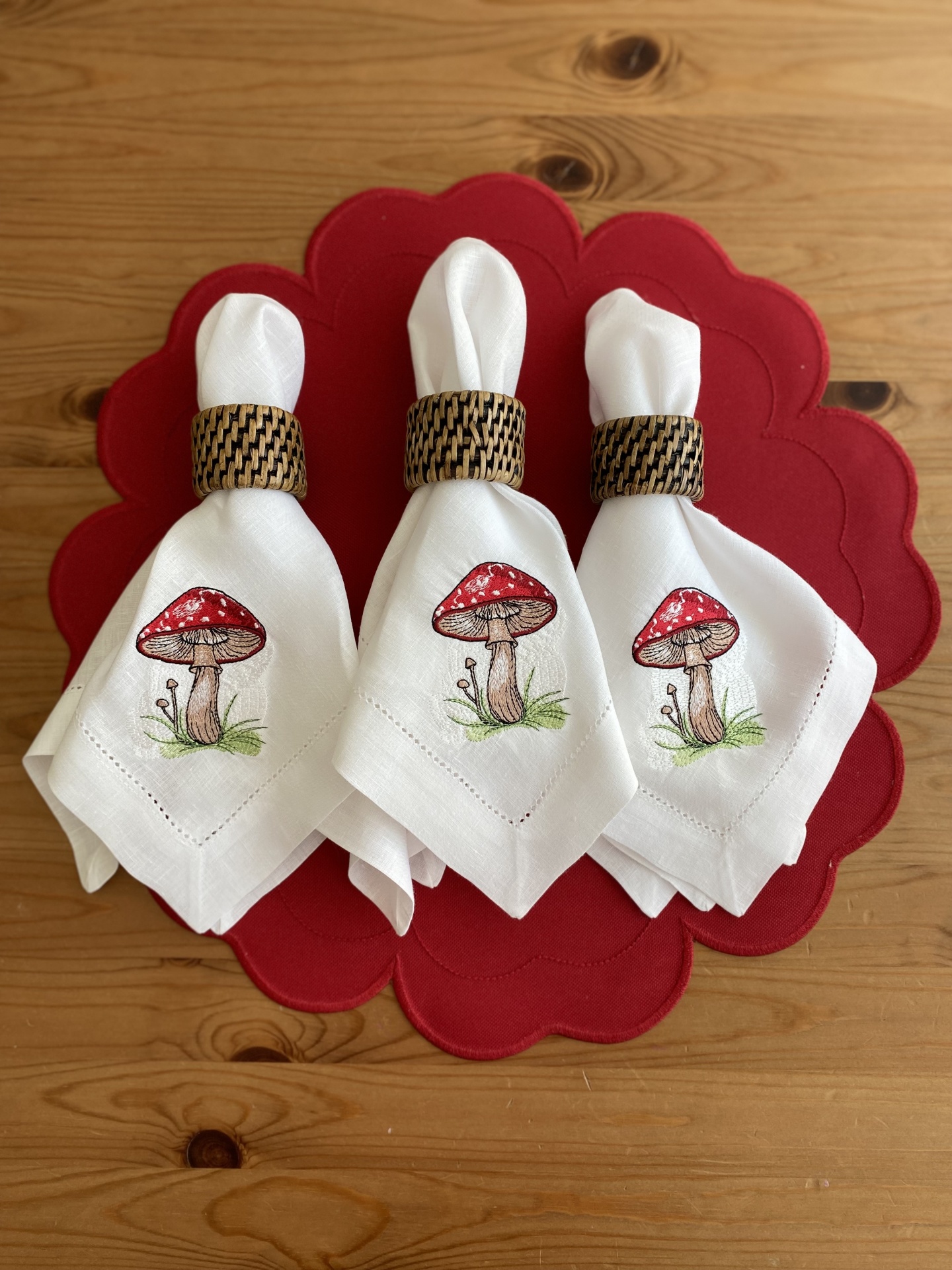 Três guardanapos brancos com cogumelos estampados e argolas em padrão castanho, sobre porta-copos vermelho, em mesa de madeira.