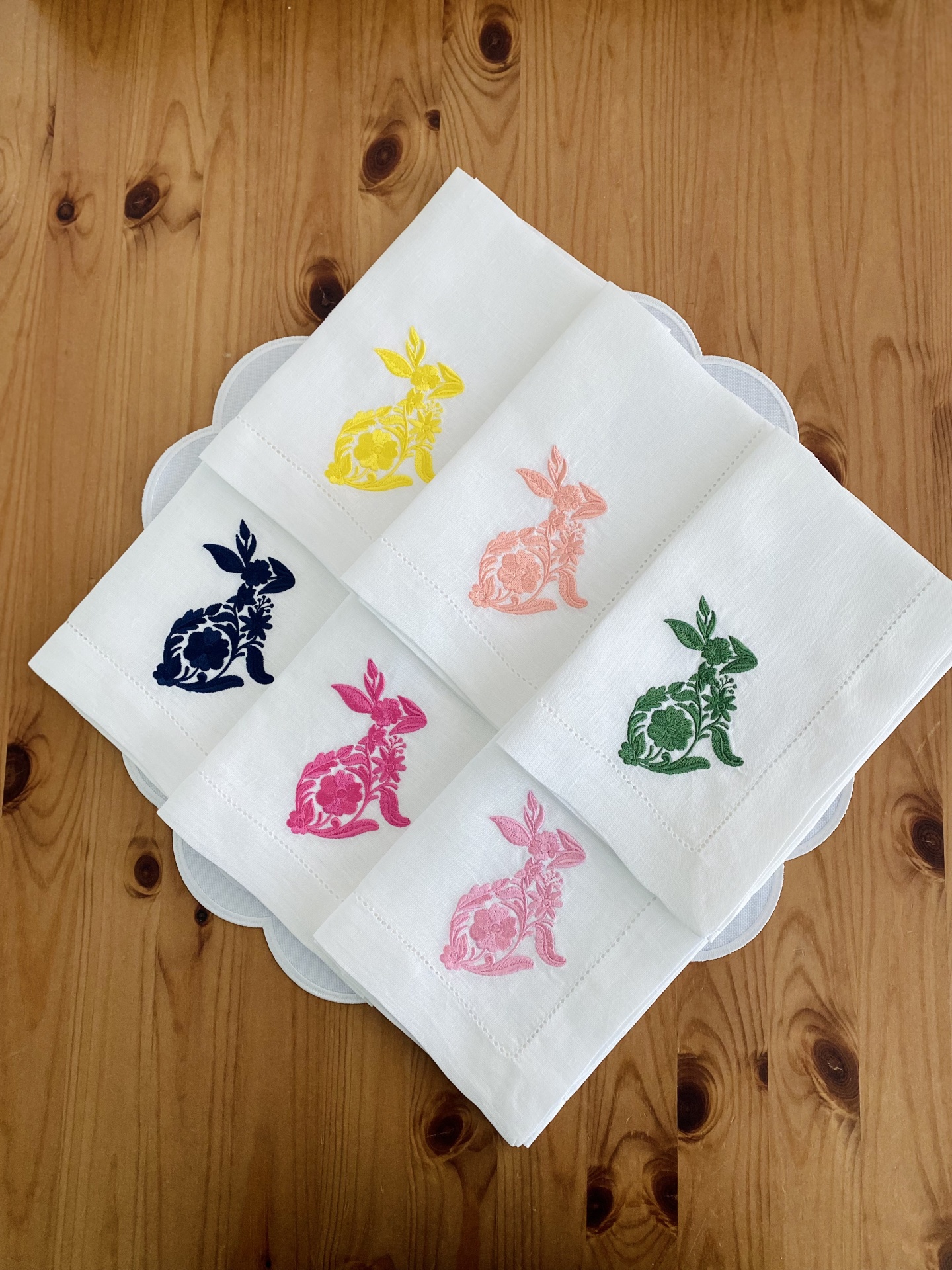 Cinco toalhas de mesa brancas com bordados coloridos de coelhos sobre prato branco e mesa de madeira.