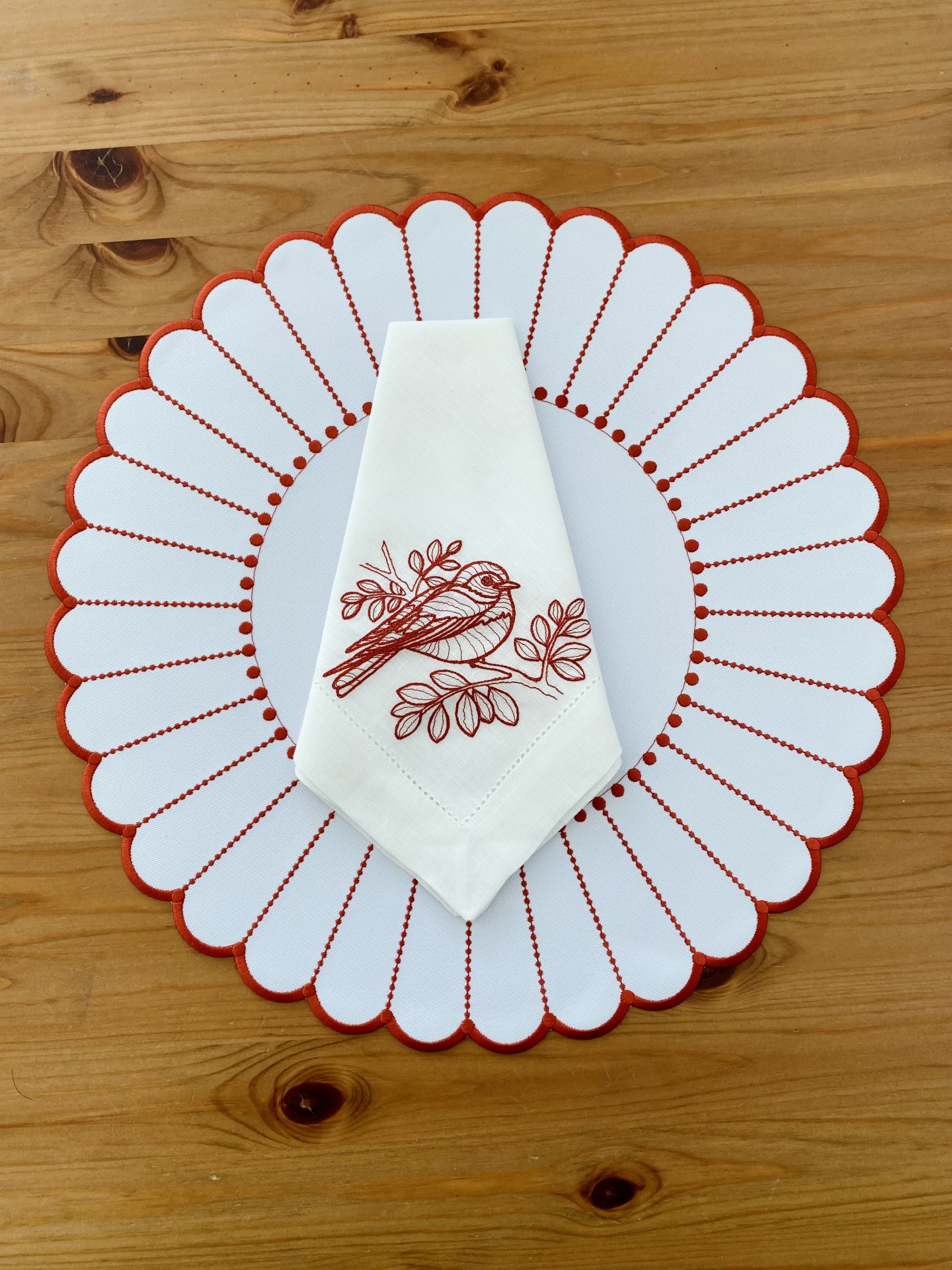 Individual branco com bordado vermelho e guardanapo branco com bordado de pássaro em tecido, sobre mesa de madeira clara