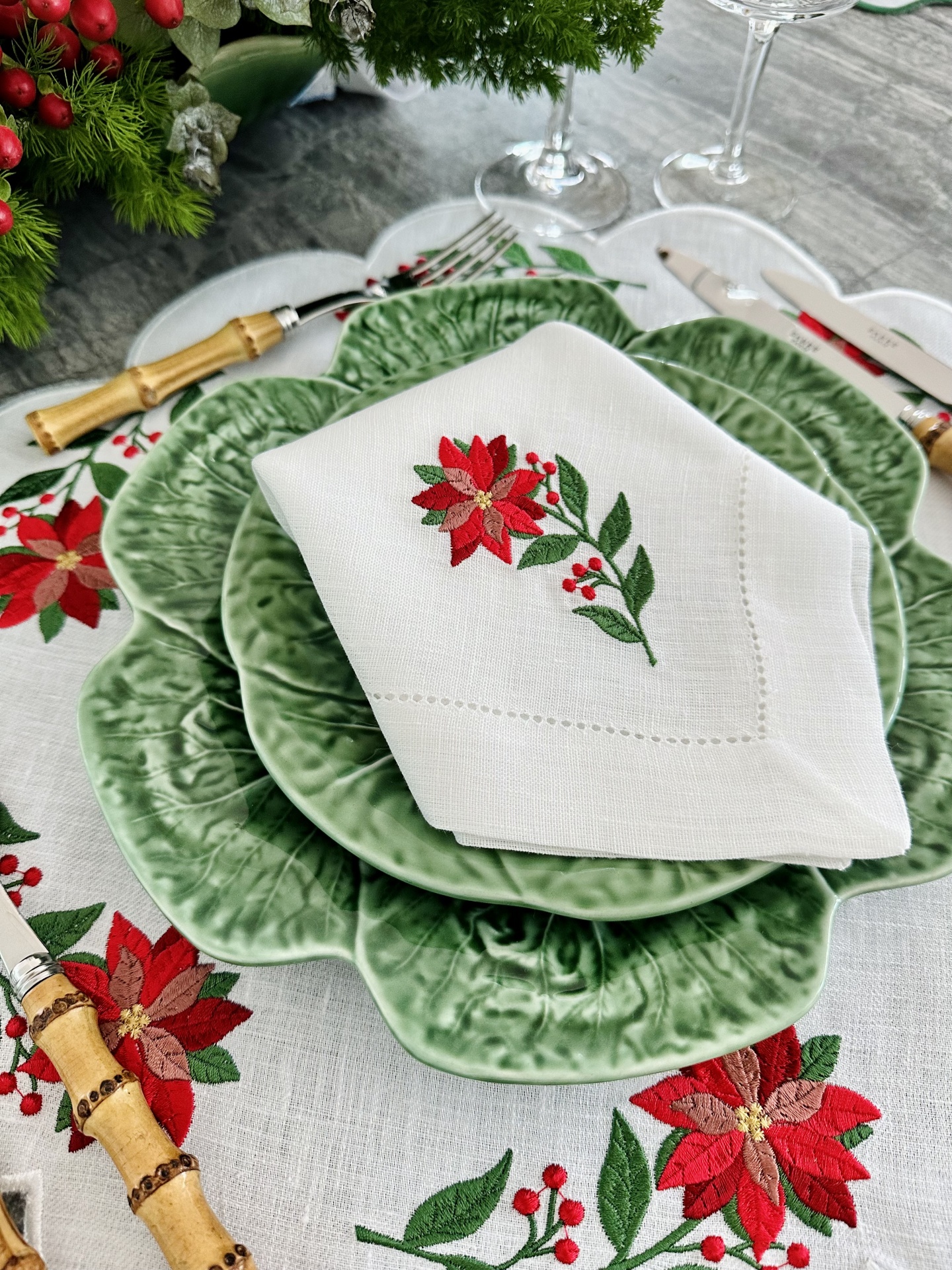 Louça verde com textura, guardanapo branco bordado com flor vermelha, talheres de madeira e prata, toalha com motivos natalícios.