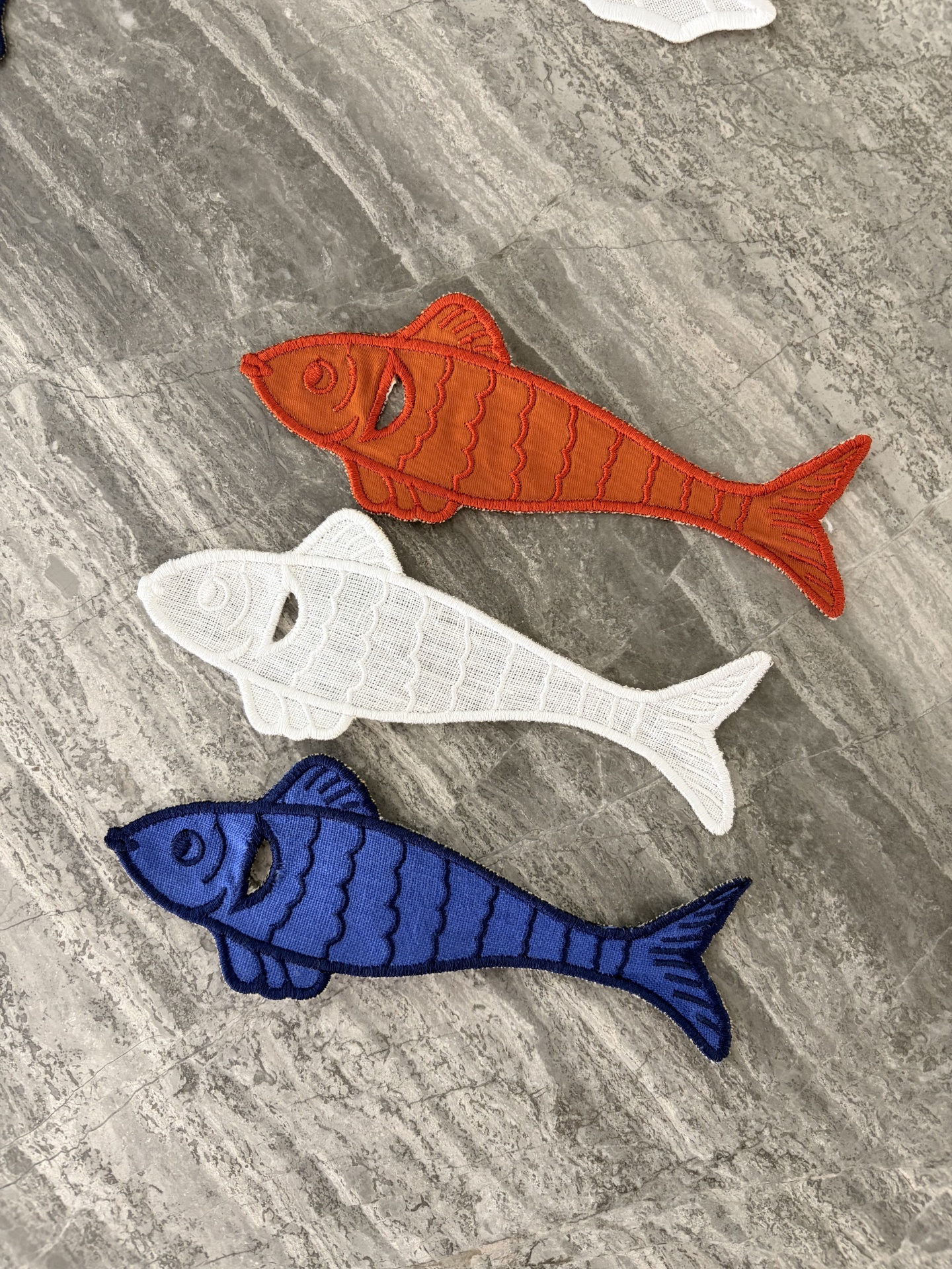Três patches bordados em forma de peixe em cores laranja, branco e azul sobre pedra cinza.