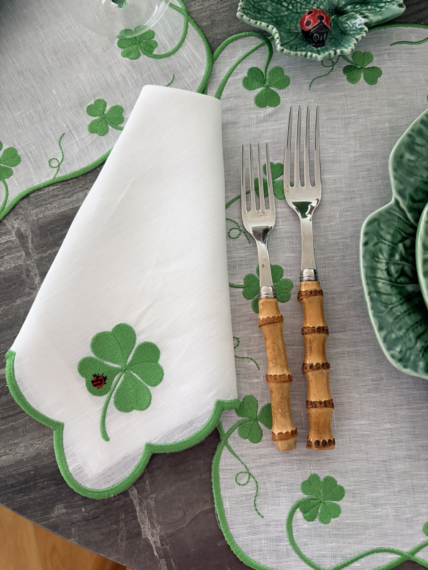 Mesa posta com guardanapo branco e bordado verde de trevo, garfos com cabo de bambu, prato verde em cerâmica e toalha com padrão de trevo