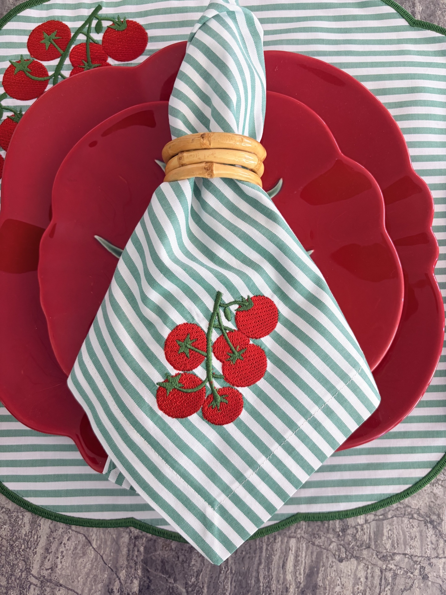 Conjunto de pratos vermelhos em forma de tomate com guardanapo às riscas verde e branco com bordado de tomates e argola de madeira.