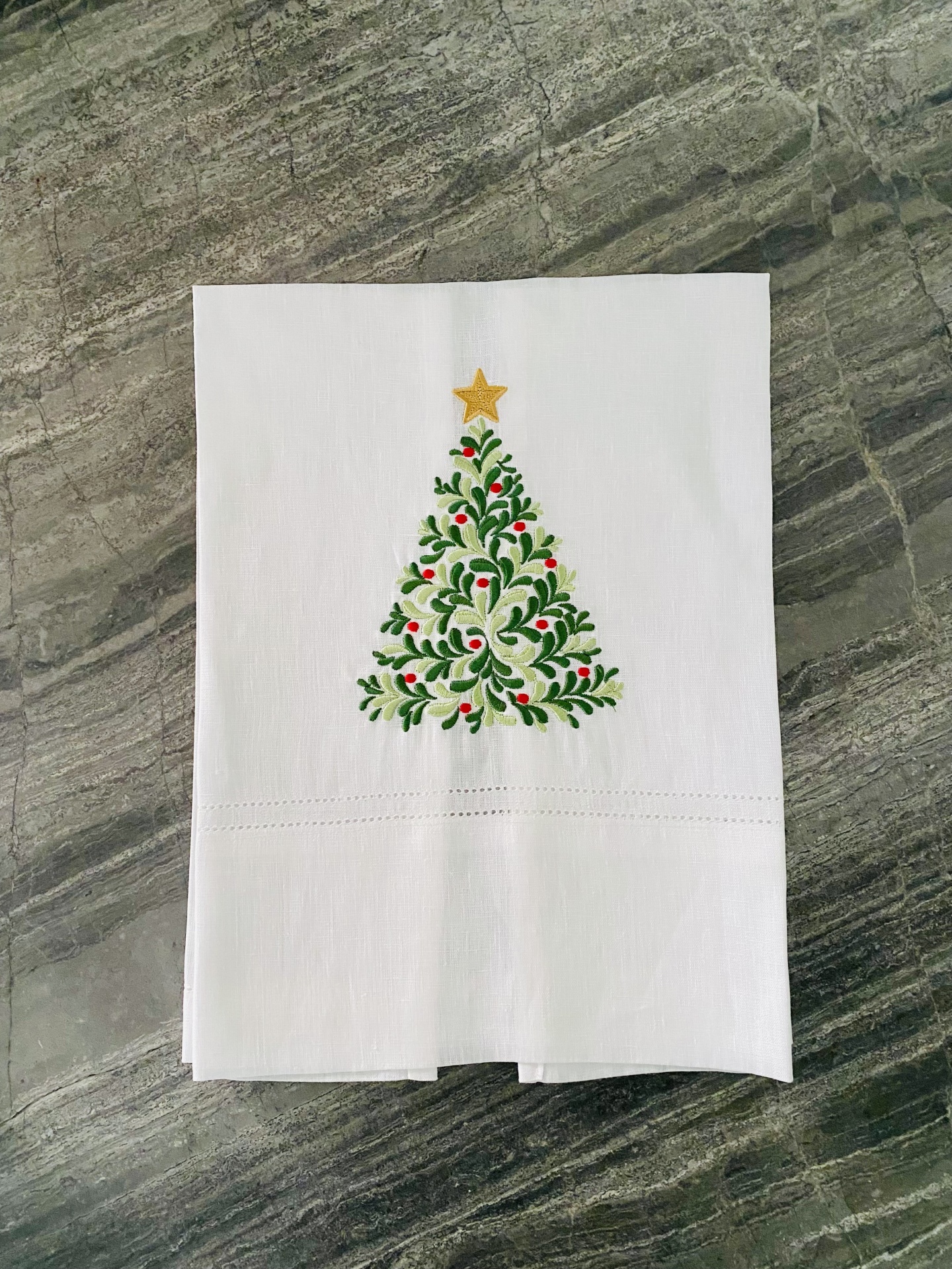 Guardanapo branco com árvore de Natal bordada em verde, vermelho e dourado sobre superfície de pedra cinzenta.