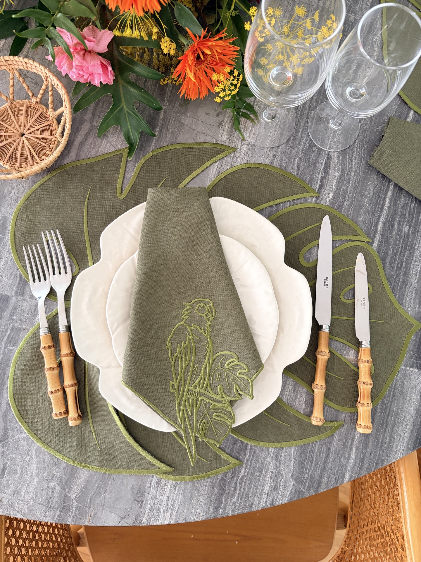 Mesa posta com toalha cinza, placemats verdes folhas, talheres bambu, pratos brancos, guardanapo verde bordado, copos vidro, flores coloridas.