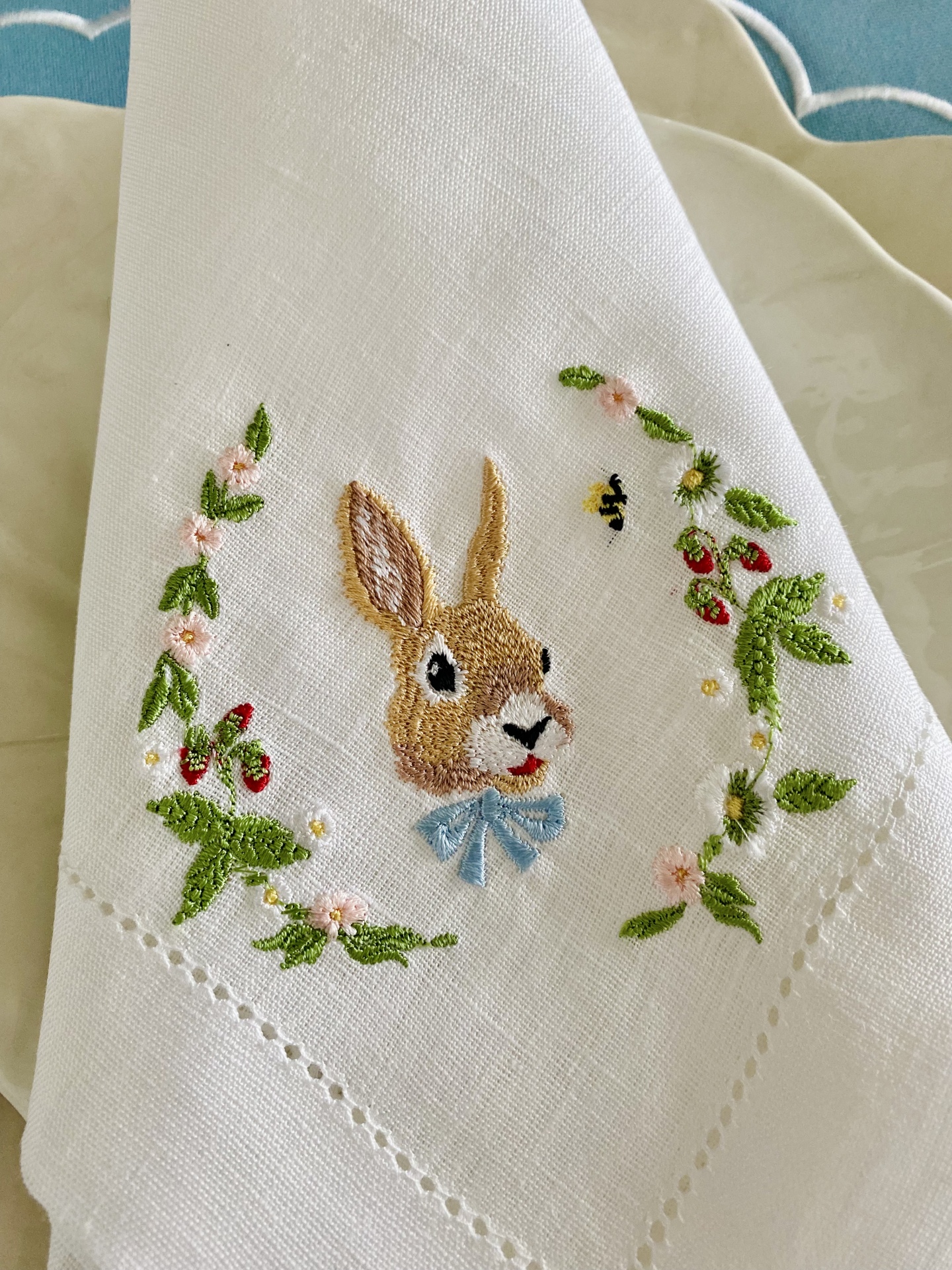 Guardanapo branco com bordado de coelho, flores e frutos vermelhos sobre prato creme