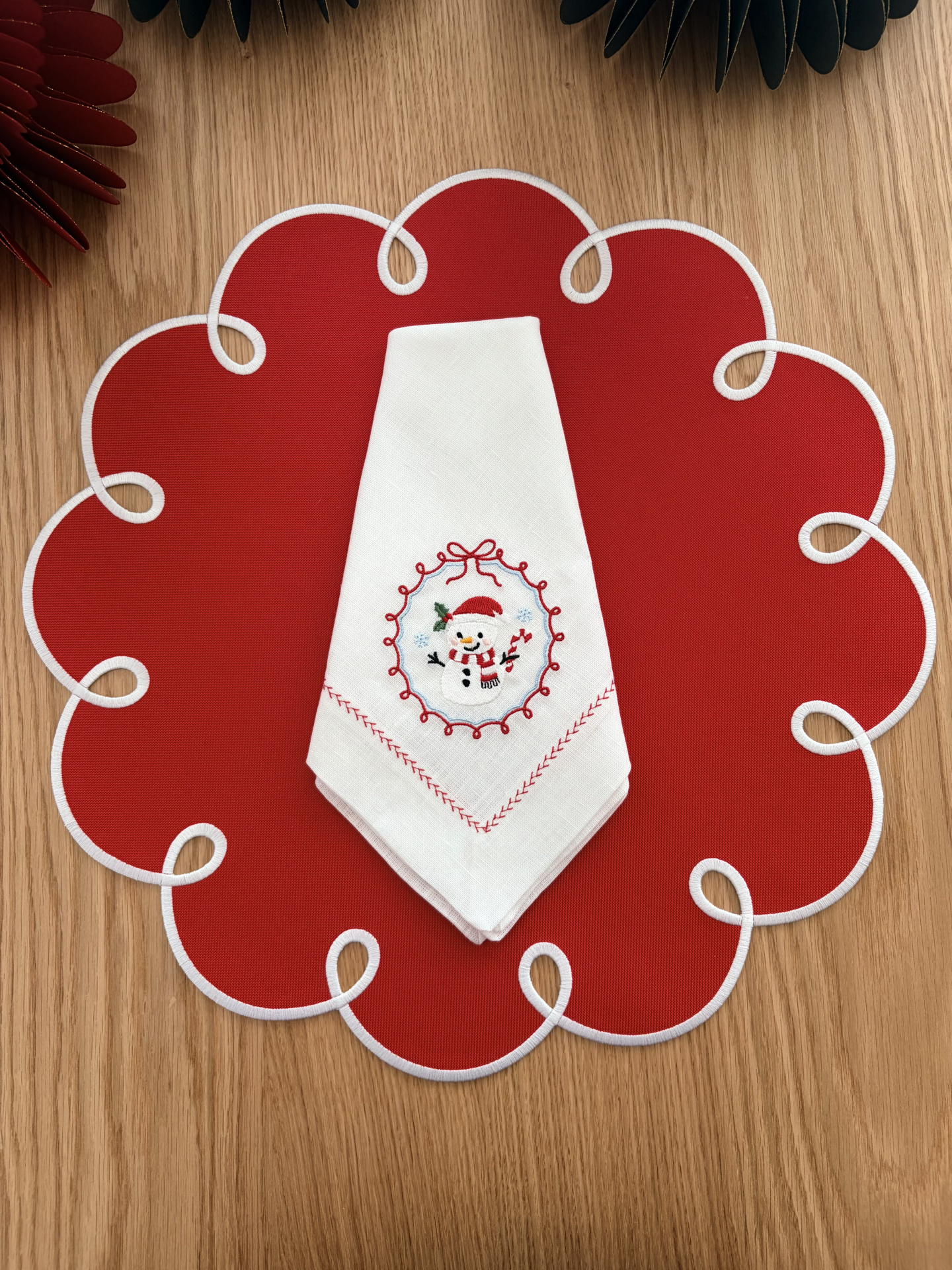 Guardanapo branco bordado com boneco de neve em jogo americano vermelho com contorno branco em mesa de madeira.