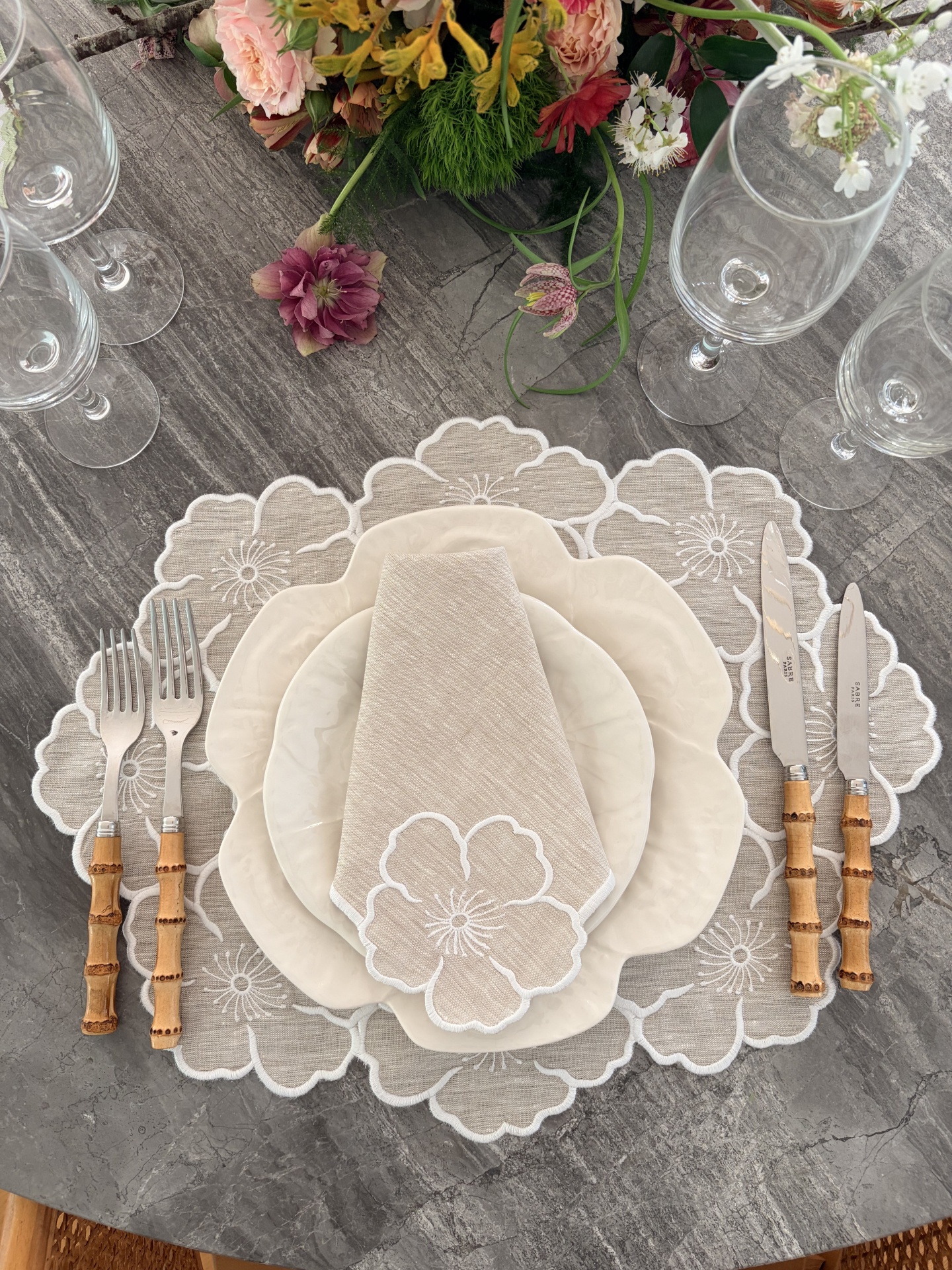 Mesa posta com pratos creme, talheres com cabos de bambu e guardanapo bege bordado com flores.