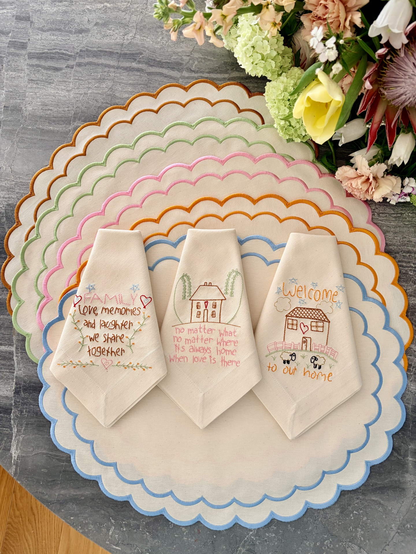 Guardanapos de tecido bordados com frases e flores sobre toalhas redondas coloridas numa mesa de pedra cinzenta ao lado de flores.