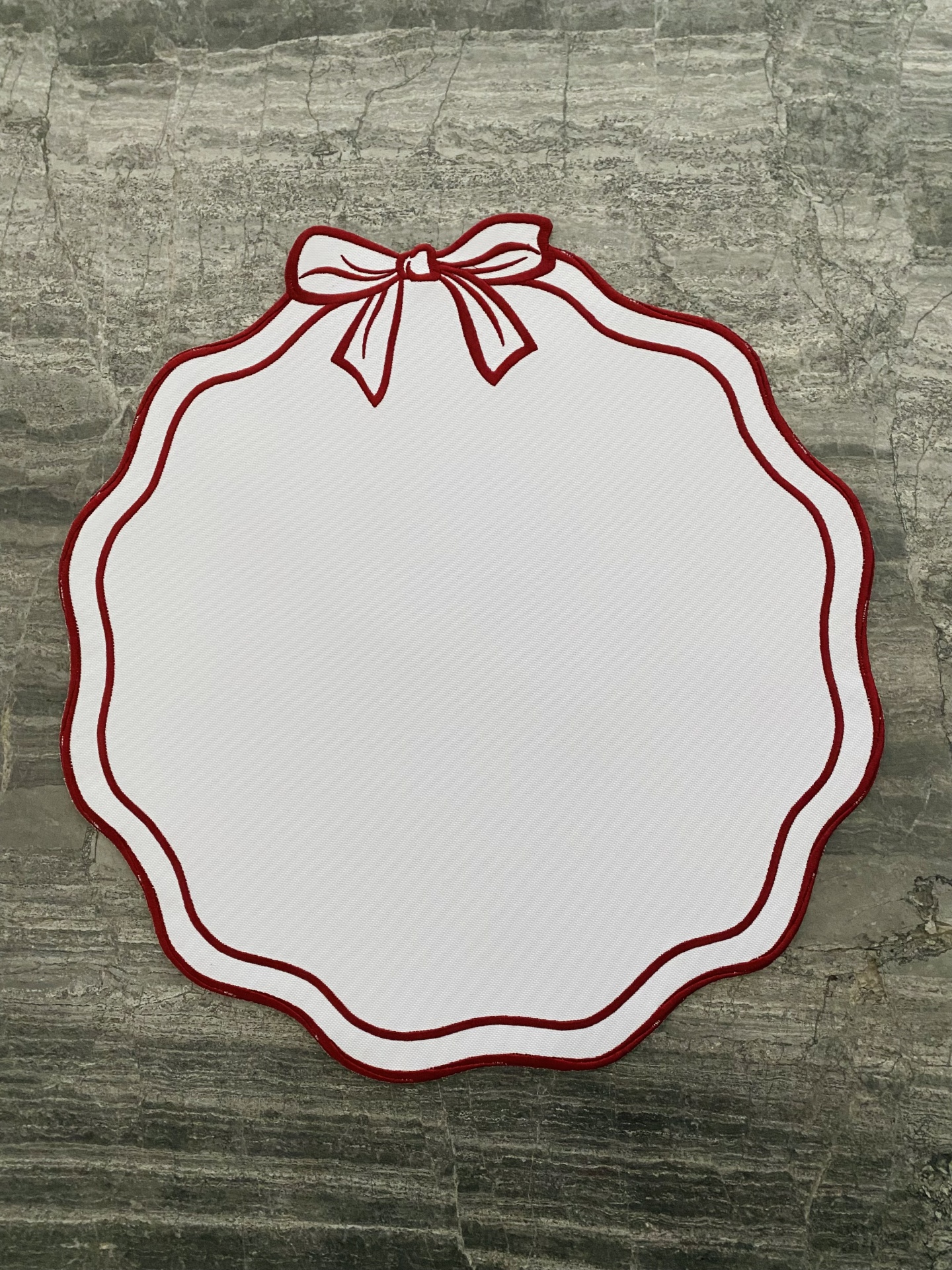 Individual de mesa branco com bordas vermelhas e desenho de laço, sobre superfície de pedra cinzenta