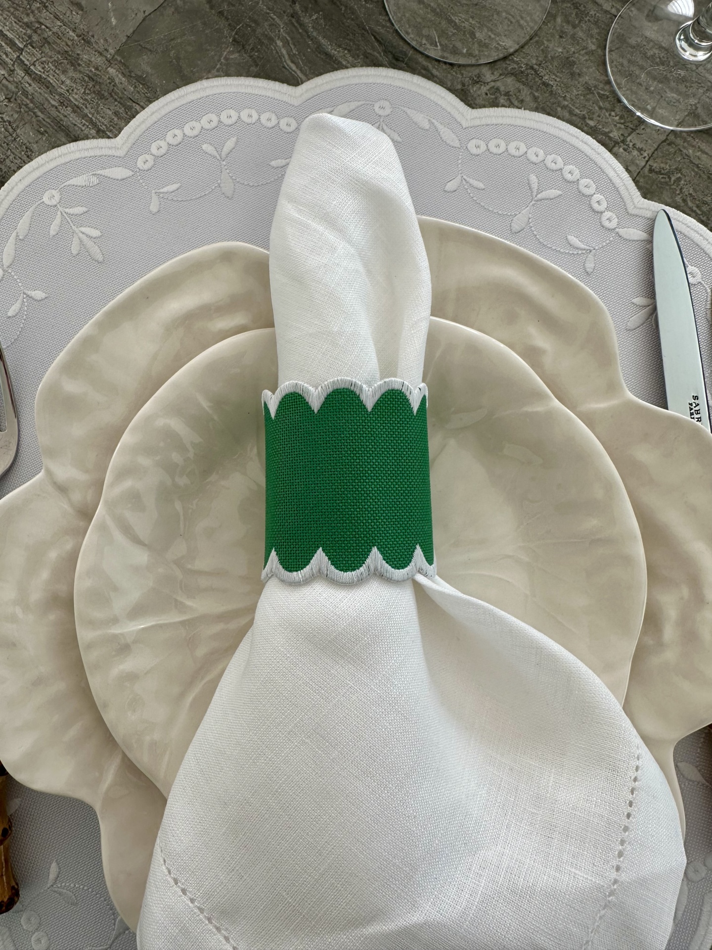 Mesa posta com pratos cerâmicos bege, guardanapo branco com anel verde e suporte cinzento bordado.