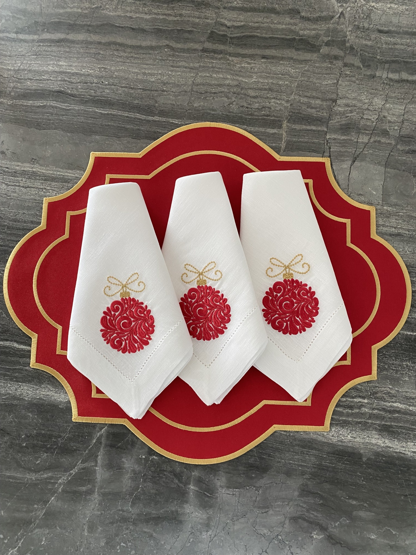 Guardanapos brancos bordados com bolas de Natal vermelhas, sobre jogo americano vermelho com borda dourada.