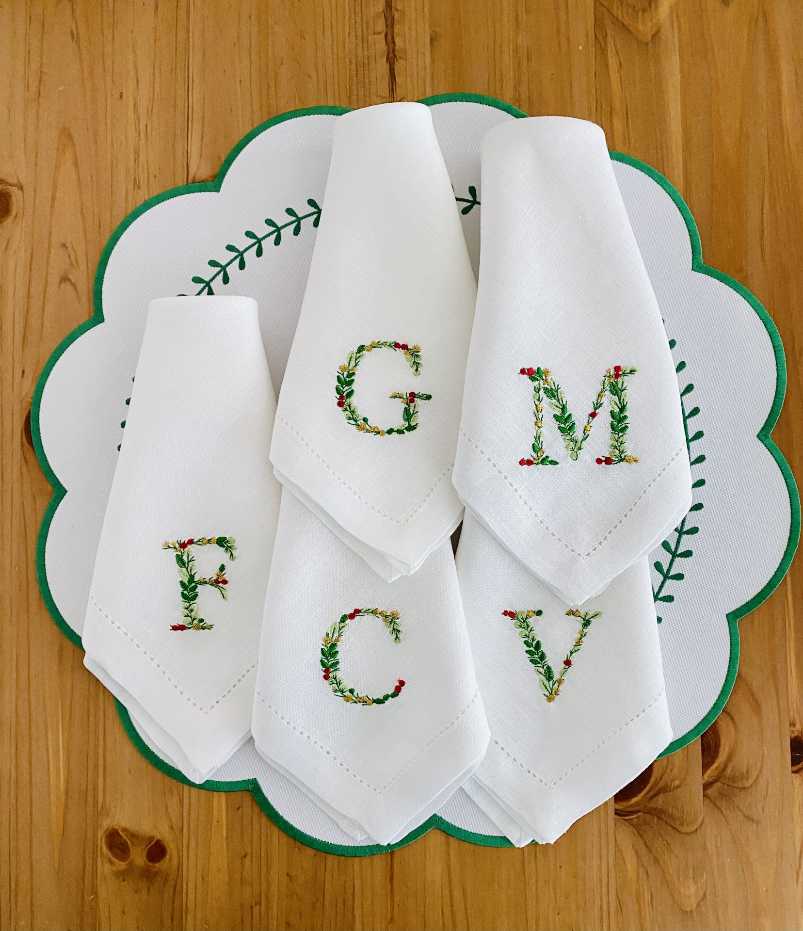 Cinco guardanapos de tecido branco bordados com letras florais sobre sousplat branco e verde, em mesa de madeira clara.