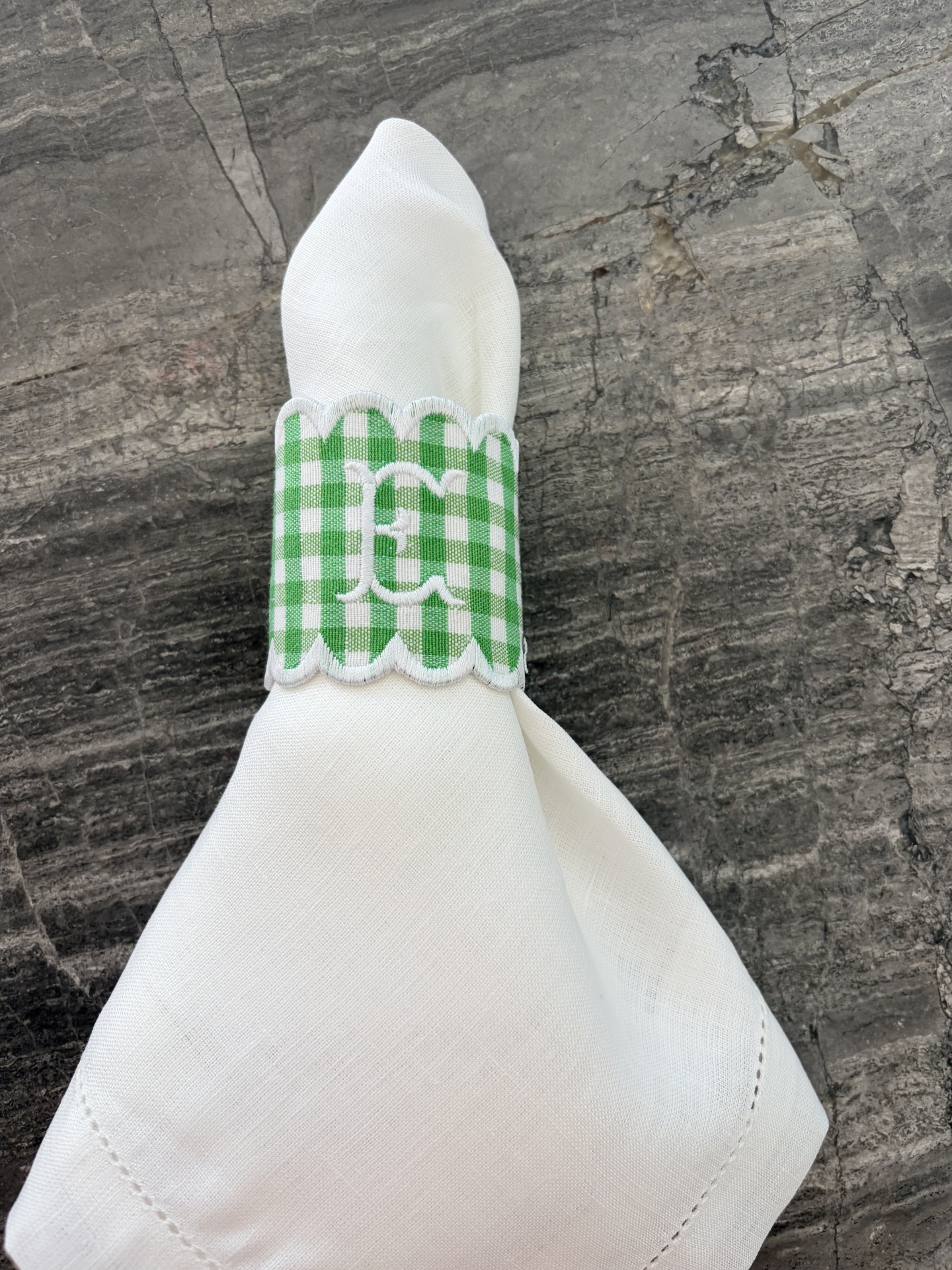Guardanapo branco com argola verde aos quadrados brancos com bordado branco, sobre pedra cinzenta.