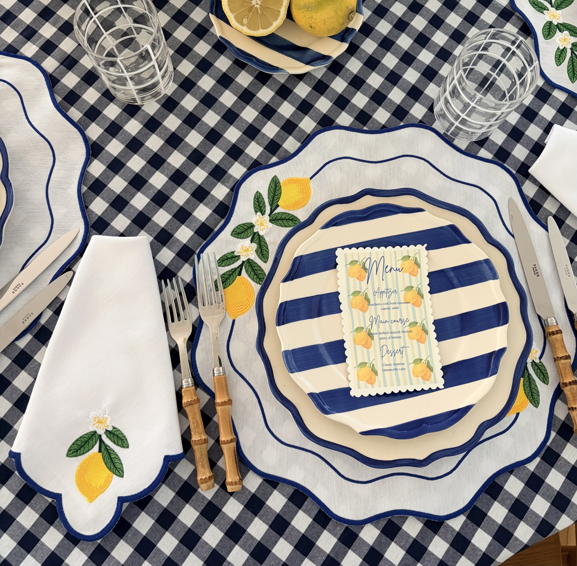 Mesa com toalha xadrez azul e branca, pratos risca azul e creme, guardanapo com bordado limão, talheres bambu, copos transparentes com padrão branco