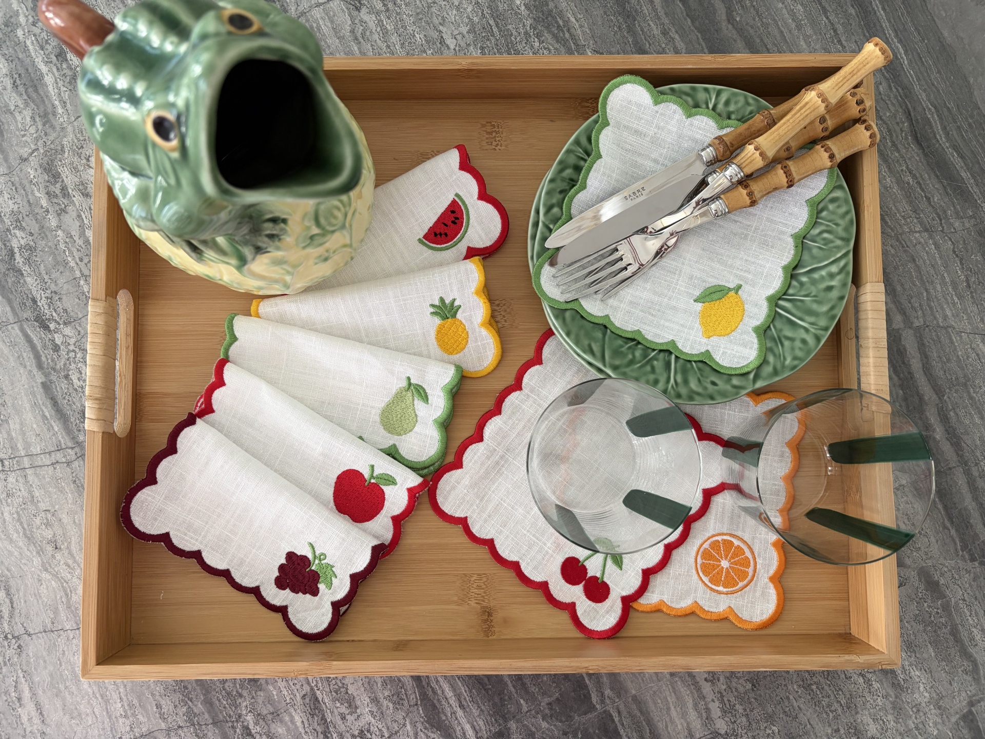 Tabuleiro com jarra cerâmica, pratos verdes, guardanapos com bordados de frutas, talheres com cabos de bambu e copos transparentes