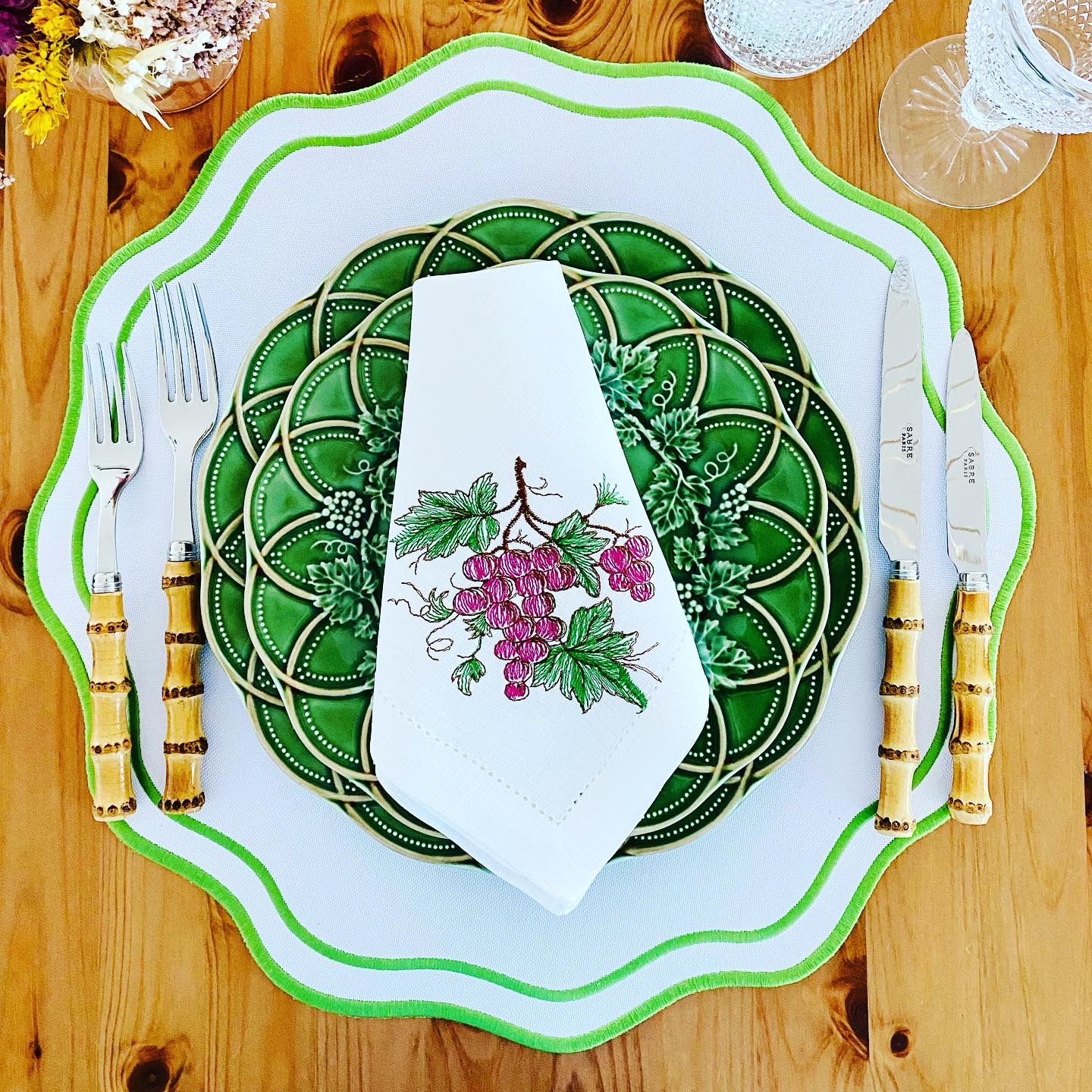 Mesa posta com pratos verdes e brancos, talheres com cabo de bambu e guardanapo bordado com uvas