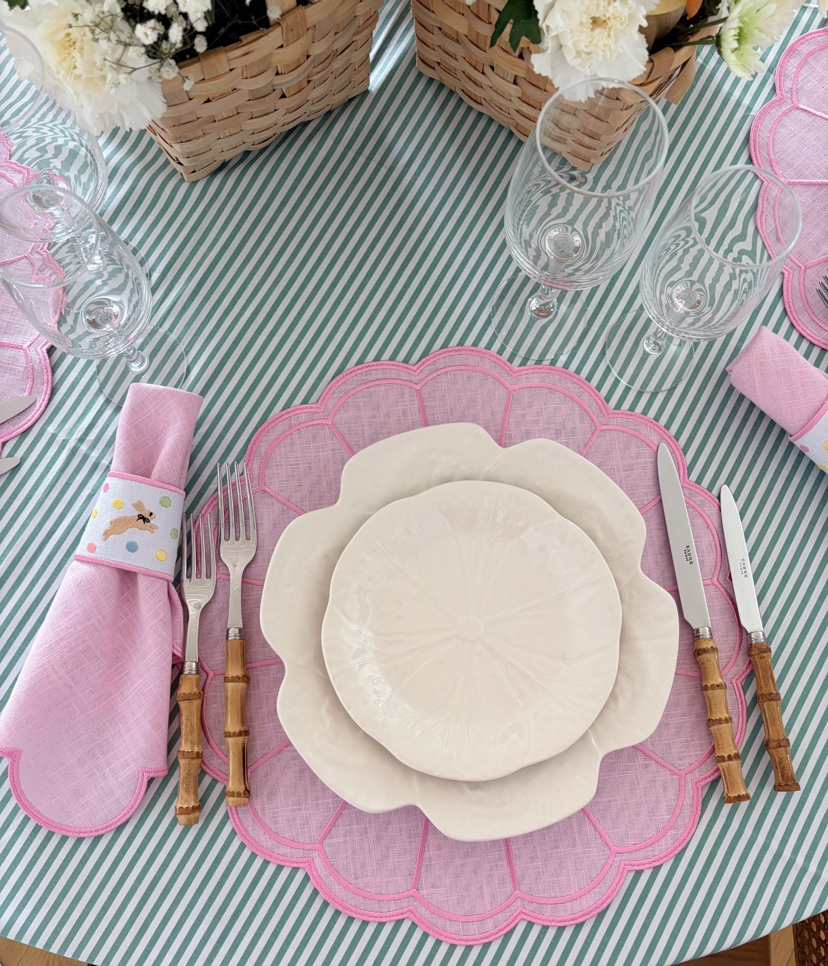 Mesa posta com pratos brancos, sousplat rosa, talheres com cabo de bambu, guardanapo rosa e toalha às riscas verdes e brancas