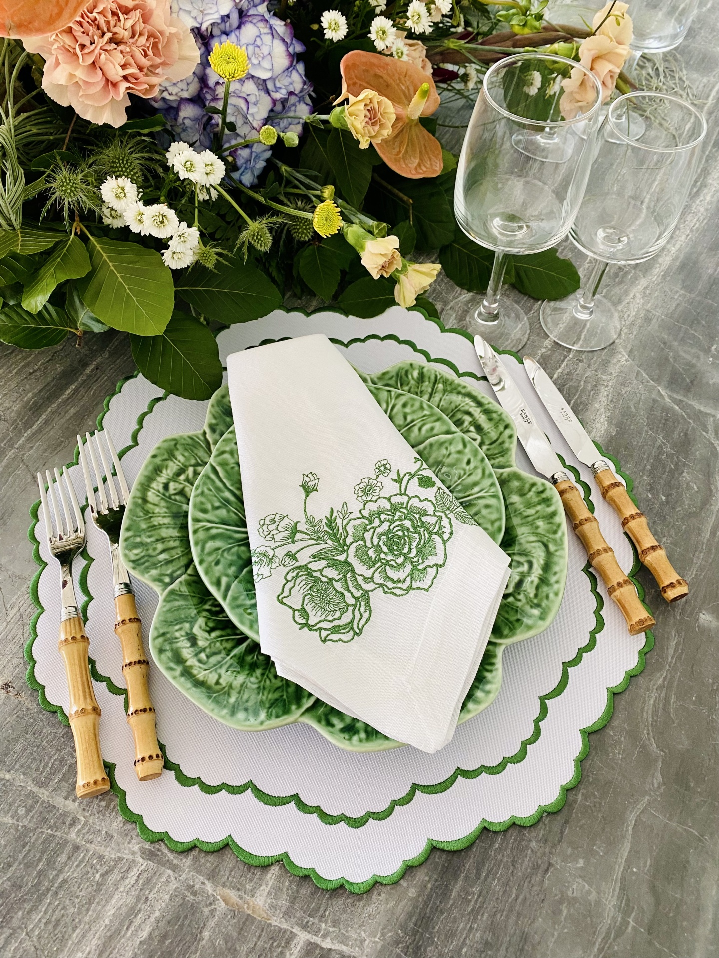 Mesa posta com pratos verdes, guardanapo branco bordado, talheres com cabo de bambu, copos de vidro e flores