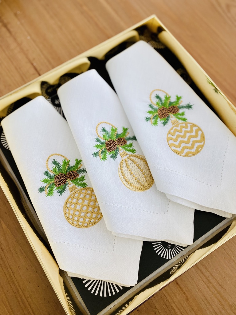 Três guardanapos brancos com bordados de bolas de Natal douradas e verdes numa caixa amarela