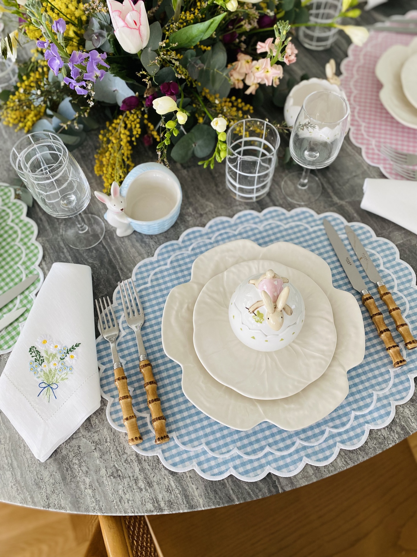 Mesa posta com pratos brancos, individual azul xadrez, talheres de madeira, guardanapo bordado, copos, caneca de coelho e arranjo floral