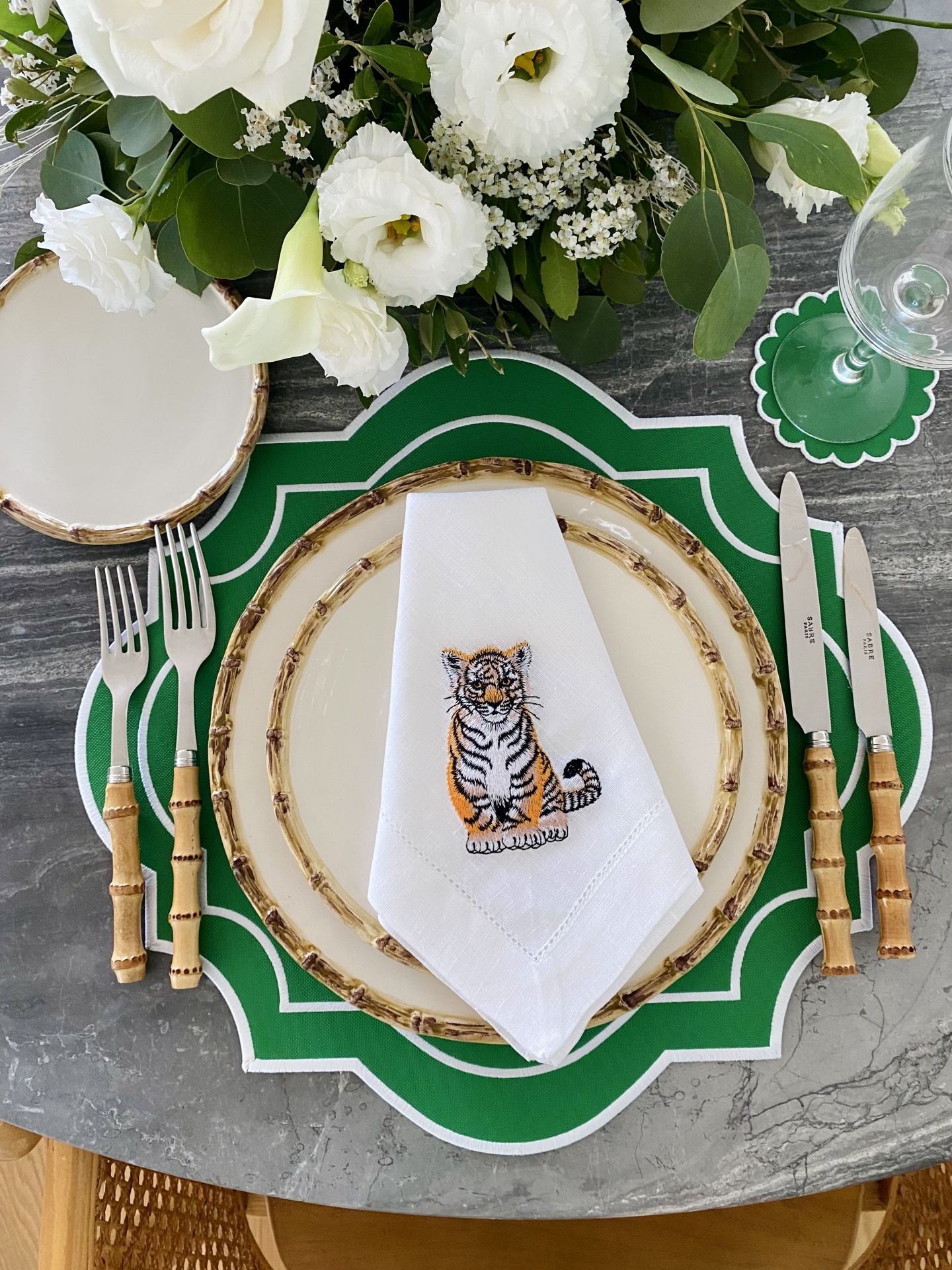 Mesa posta com pratos com borda de bambu, talheres com cabos de bambu, guardanapo branco bordado com tigre, base de prato verde e flores brancas no centro