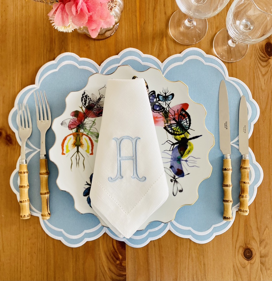 Mesa posta com prato de borboletas, guardanapo bordado, talheres com cabos imitação bambu, copos e flor rosa