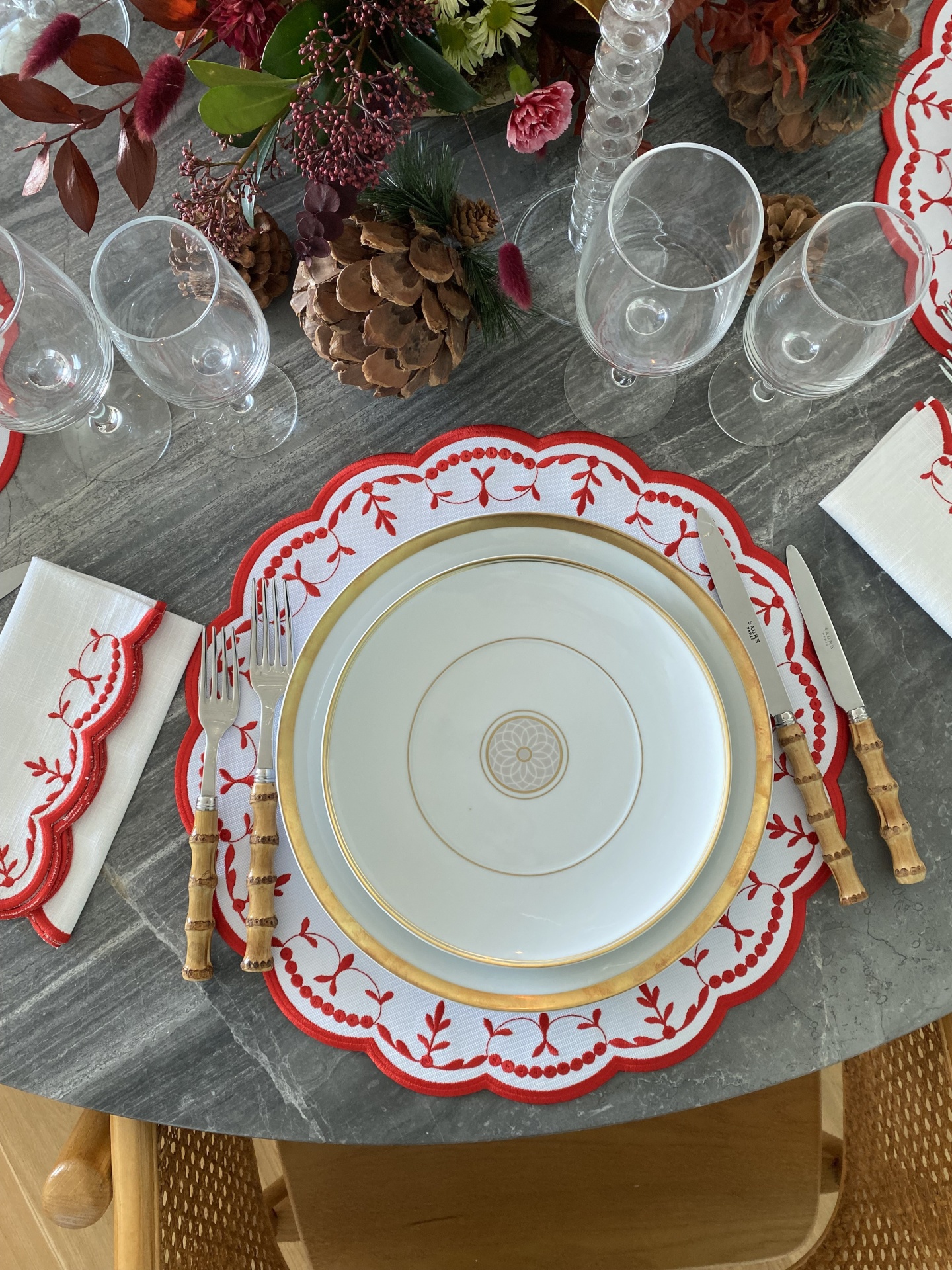 Mesa com serviço de jantar branco e dourado, base decorativa vermelha, talheres com cabo de bambu, copos transparentes e decoração floral seca