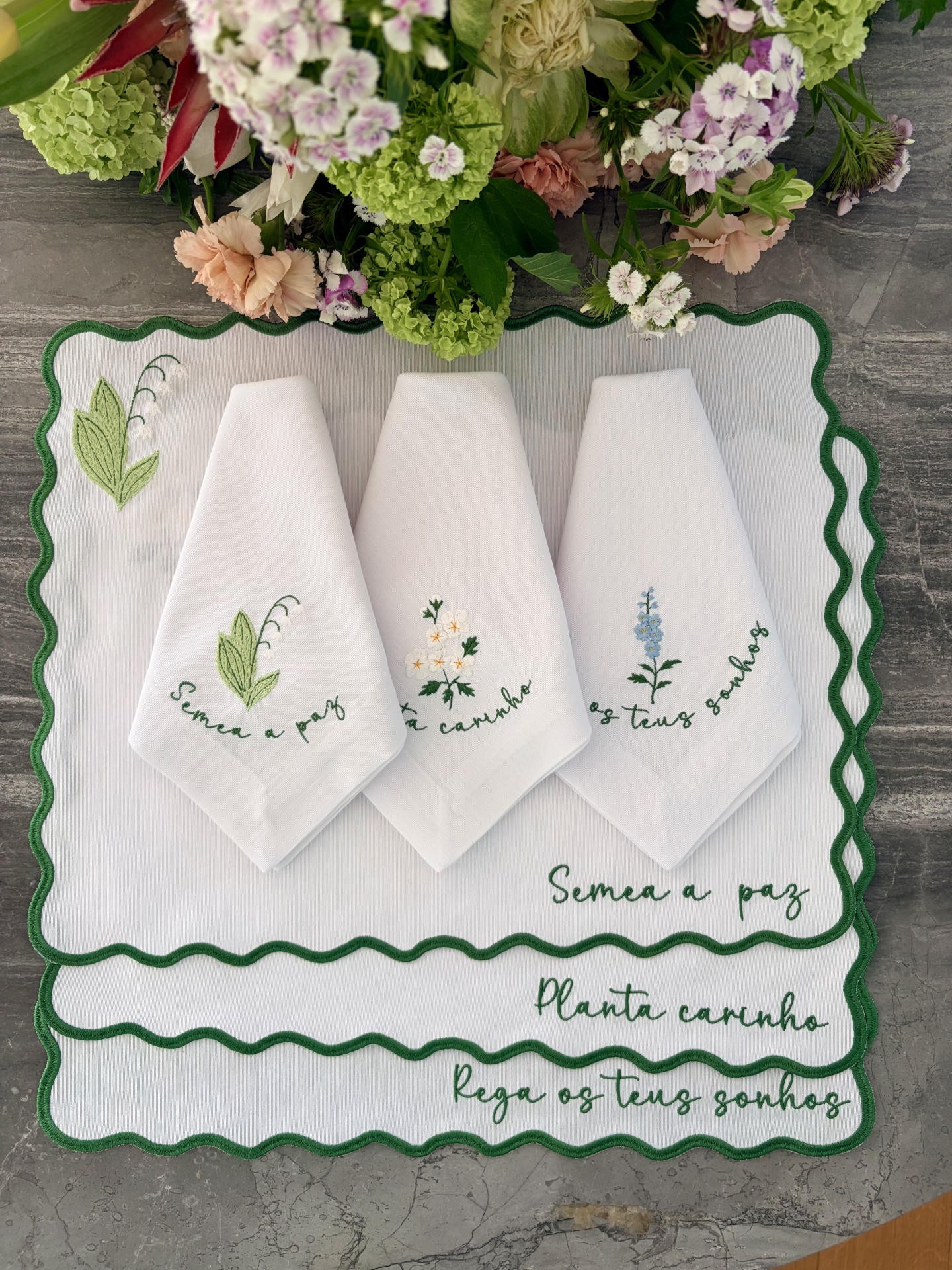 Jogo de toalhas de pano brancas com bordados de flores e texto verde, sobre toalha maior com bordado e acabamento ondulado verde.