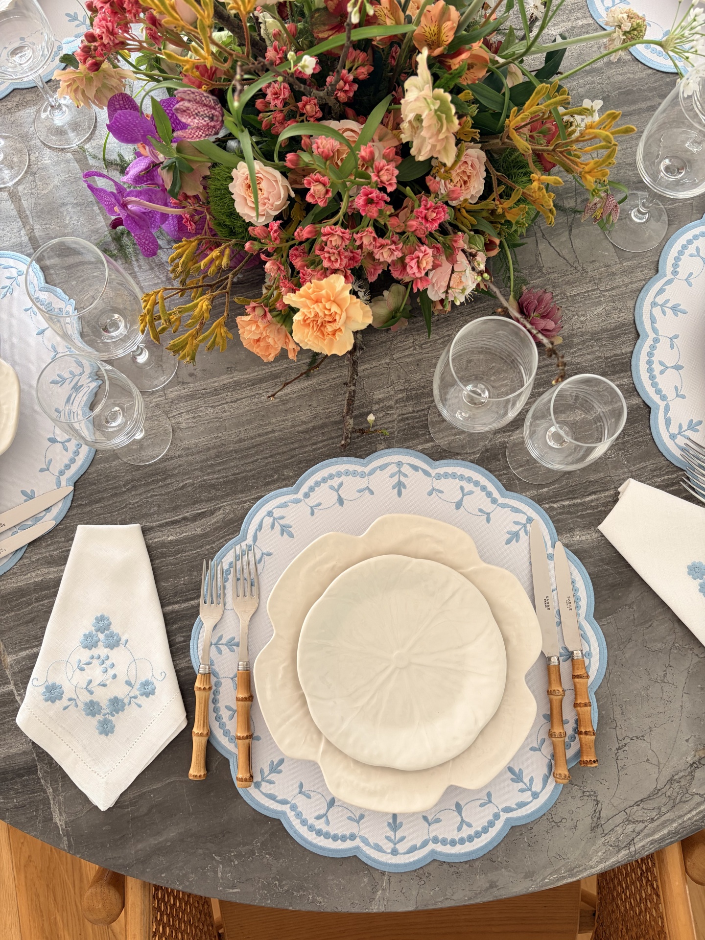 Mesa posta com louça branca em formato floral, talheres com cabo de bambu, guardanapos bordados e arranjo de flores coloridas