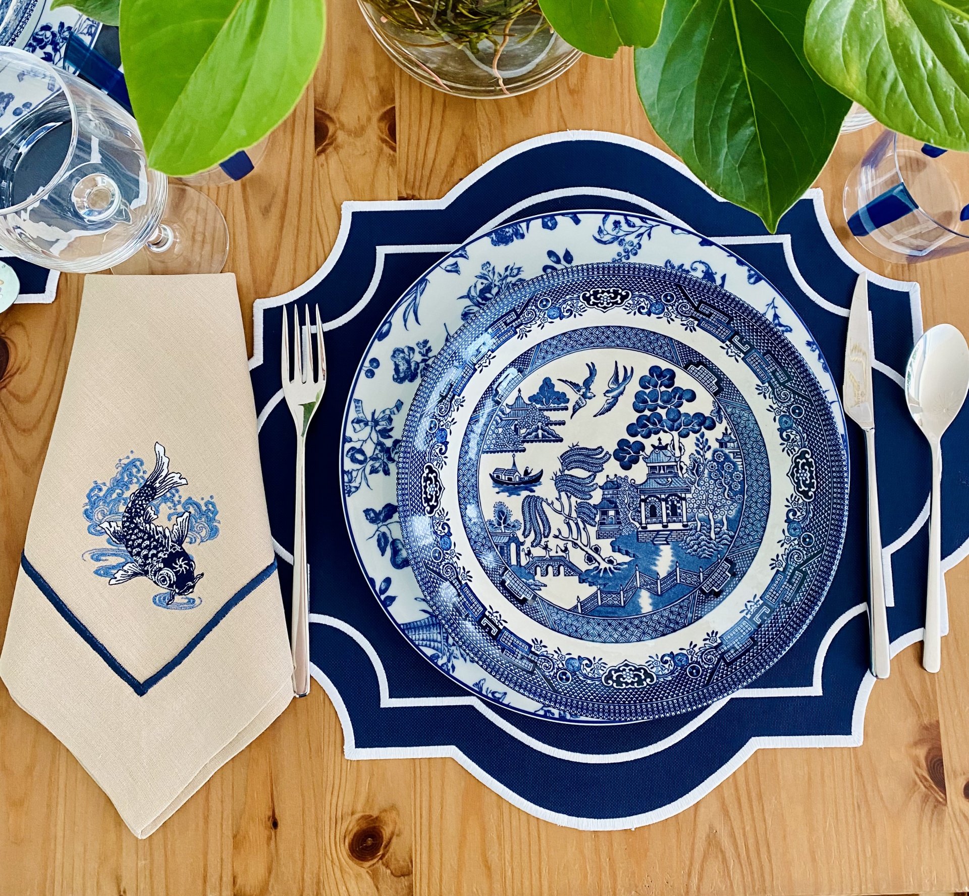 Prato de porcelana azul e branco com padrão oriental, guardanapo bege com bordado azul, talheres e copos sobre mesa de madeira com folhas verdes