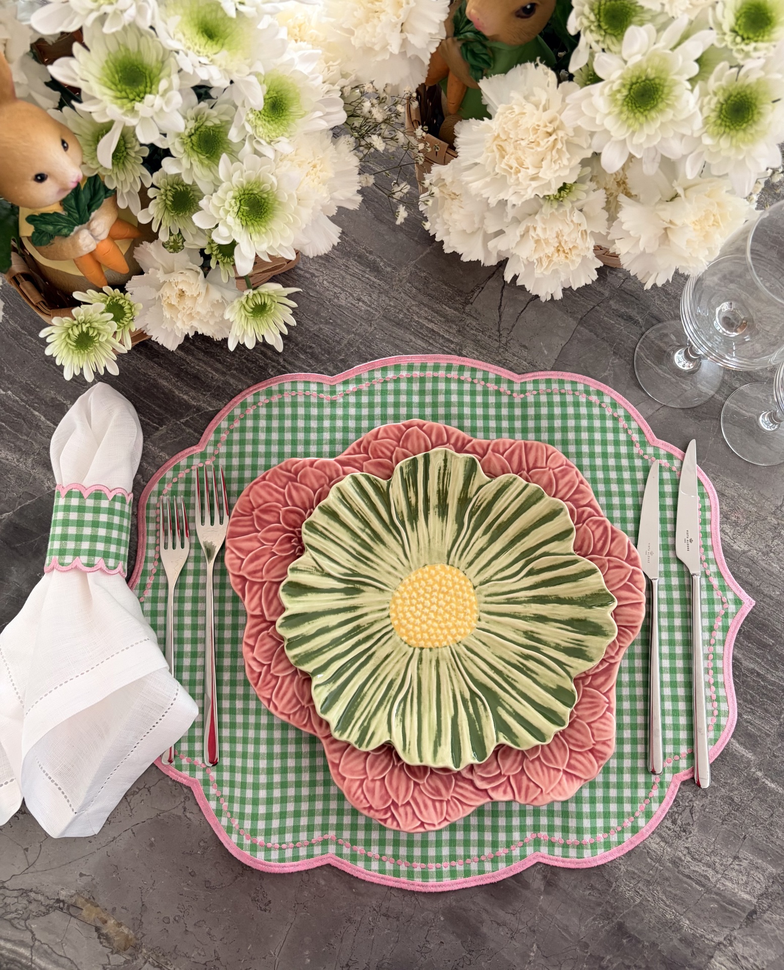 Mesa posta com pratos em forma de flor, toalhete xadrez verde e branco, talheres, guardanapo branco e flores brancas.