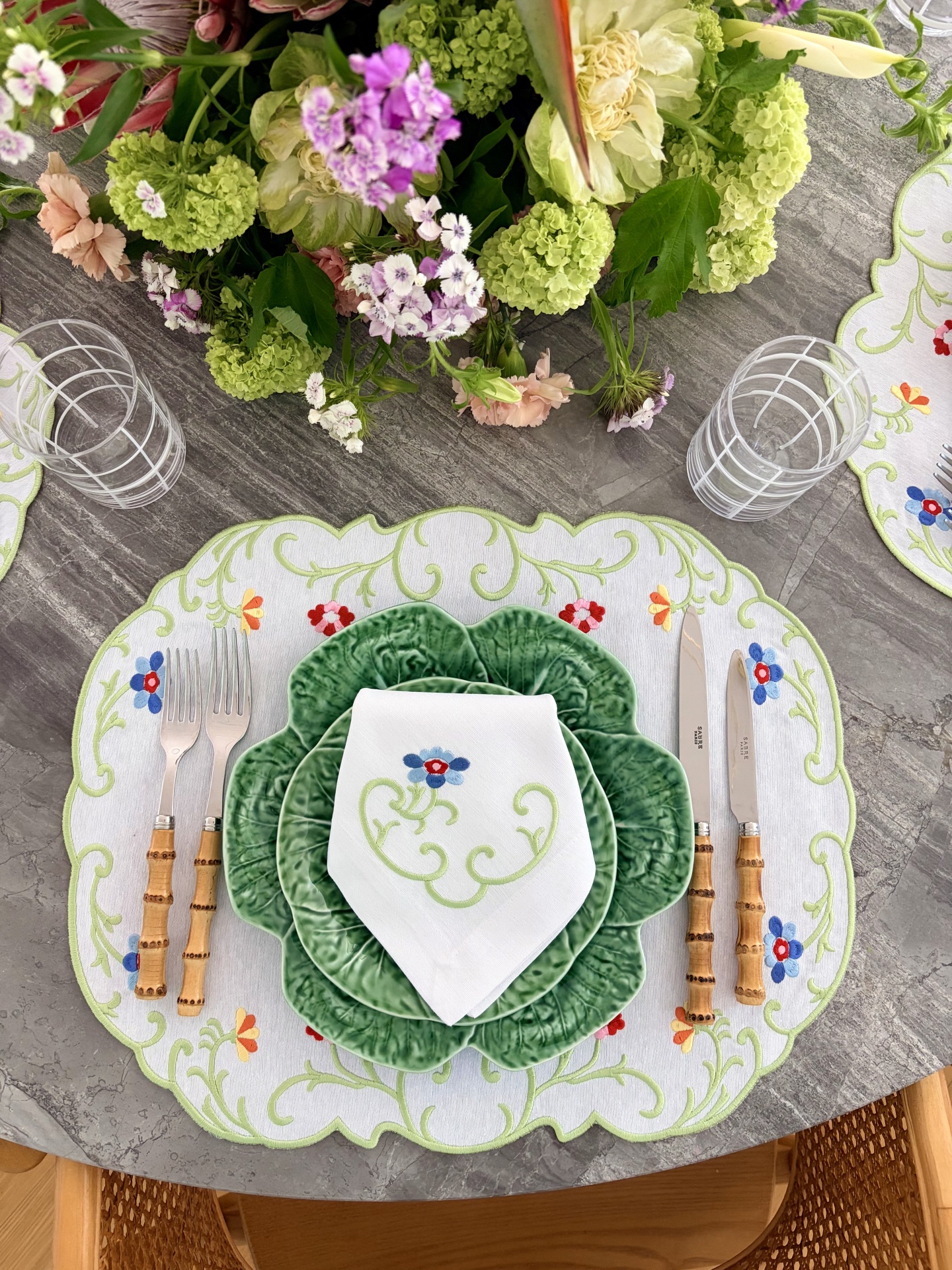 Mesa posta com pratos verdes em forma de folhas, guardanapo branco bordado e talheres com cabo amarelo sobre toalha branca bordada e arranjo floral colorido.