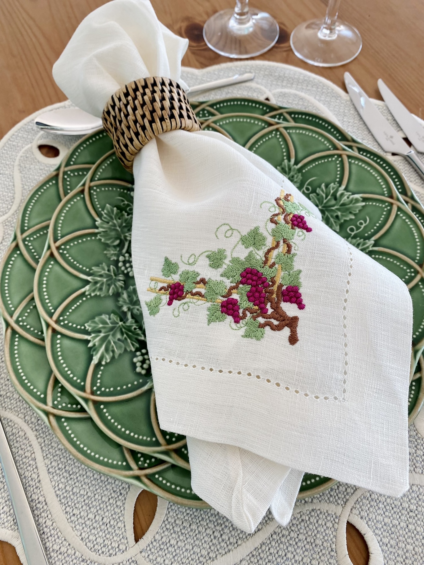 Guardanapo branco bordado com ramo de uvas sobre prato verde cerâmico, guardanapo preso por argola de madeira trançada numa mesa de madeira clara