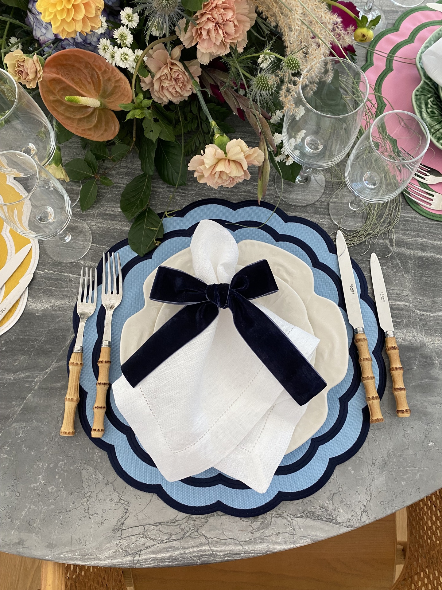 Mesa posta com base azul, prato branco, guardanapo branco com laço azul, talheres com cabo bambu e arranjo floral