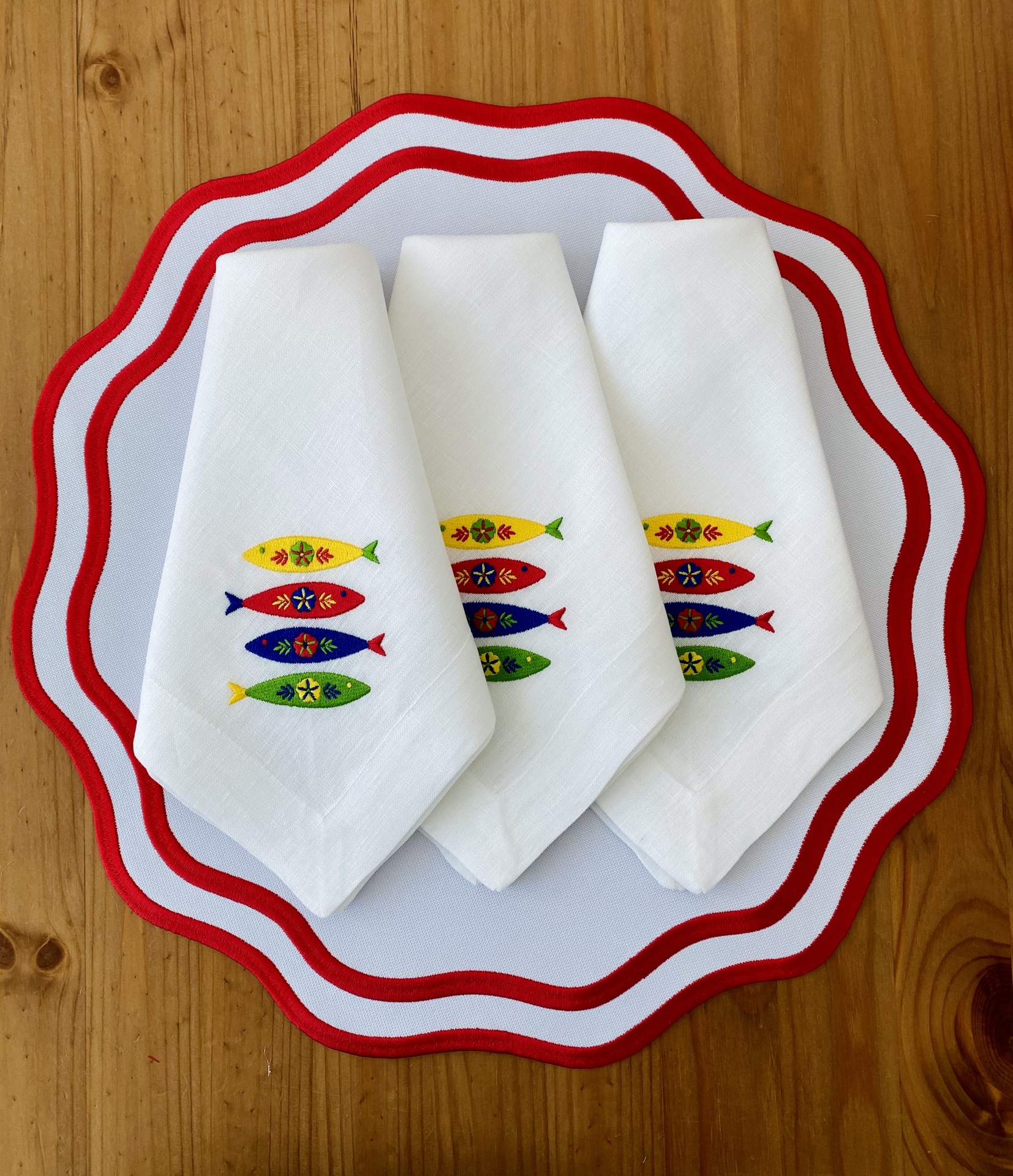 Guardanapos brancos com bordado colorido de peixes sobre tabuleiro branco com borda vermelha