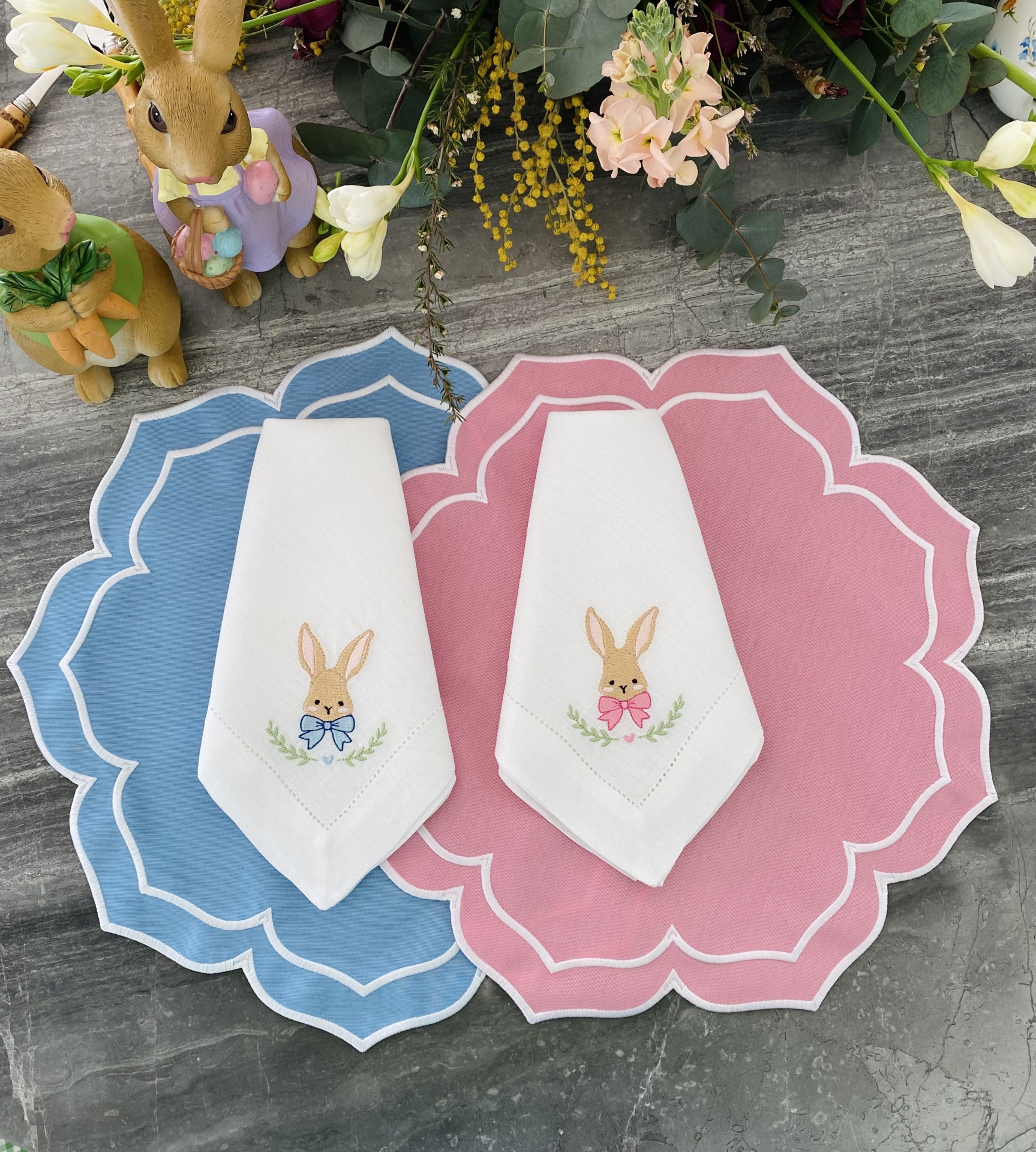 Duas peças de tecido branco com bordado de coelhos, sobre suportes de mesa rosa e azul, com decoração primaveril