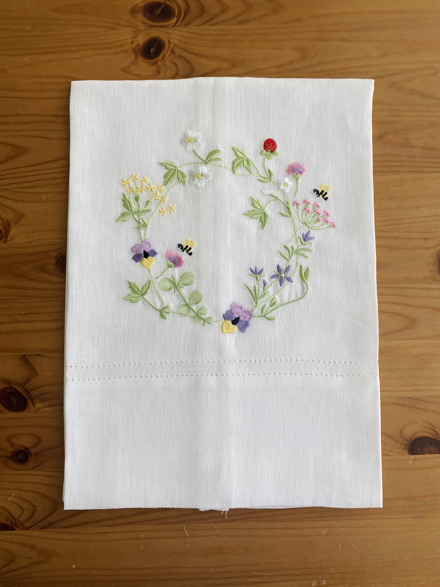 Guardanapo branco com bordado colorido de flores e abelhas sobre mesa de madeira