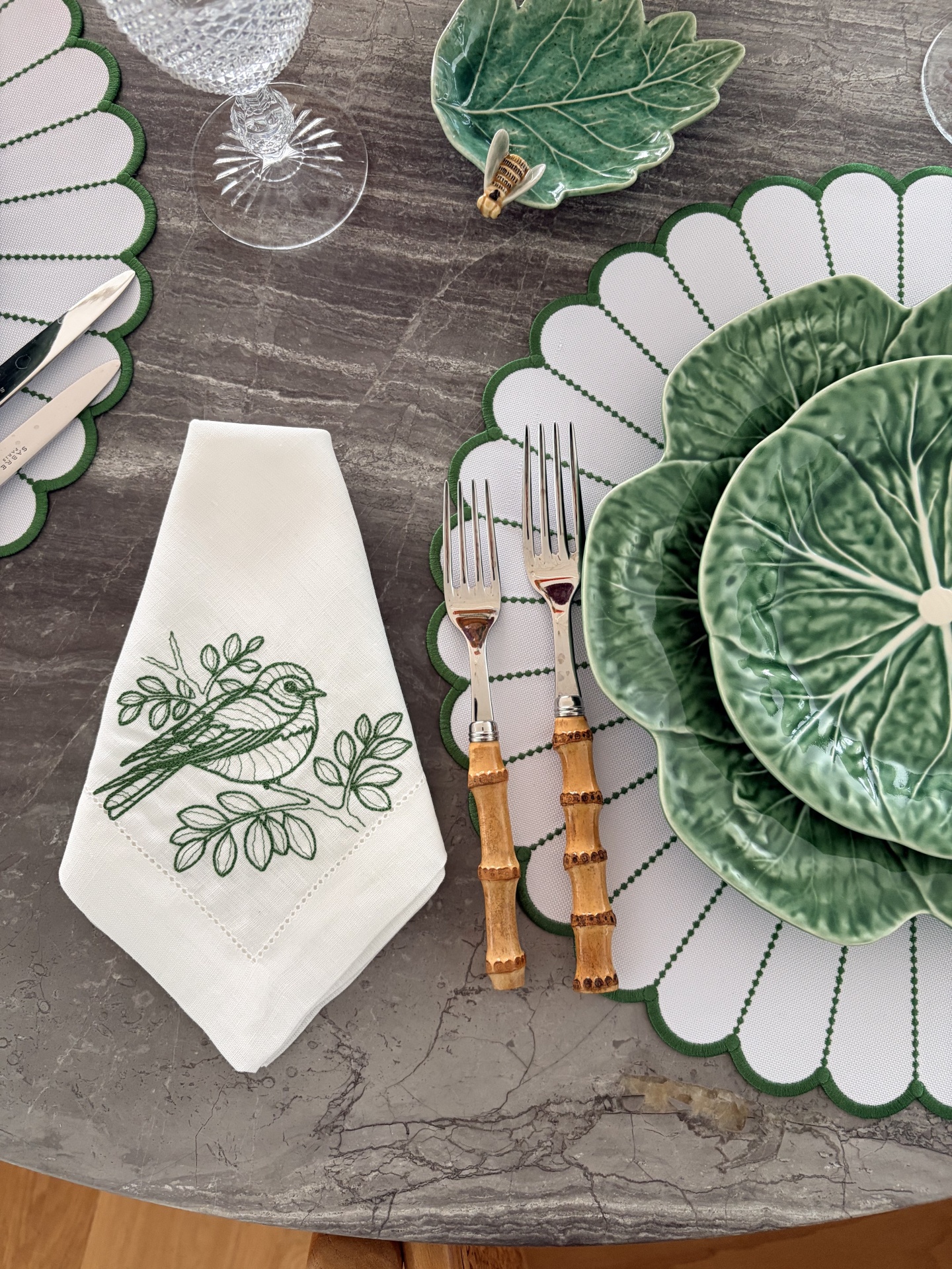 Mesa de jantar com loiça verde em forma de folha, talheres com cabo de bambu, e guardanapo branco bordado com pássaro verde